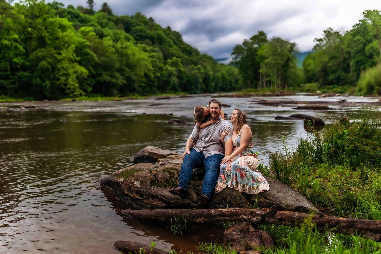 Couple sitting on rocks by a scenic river surrounded by lush green trees and mountains on a cloudy day.