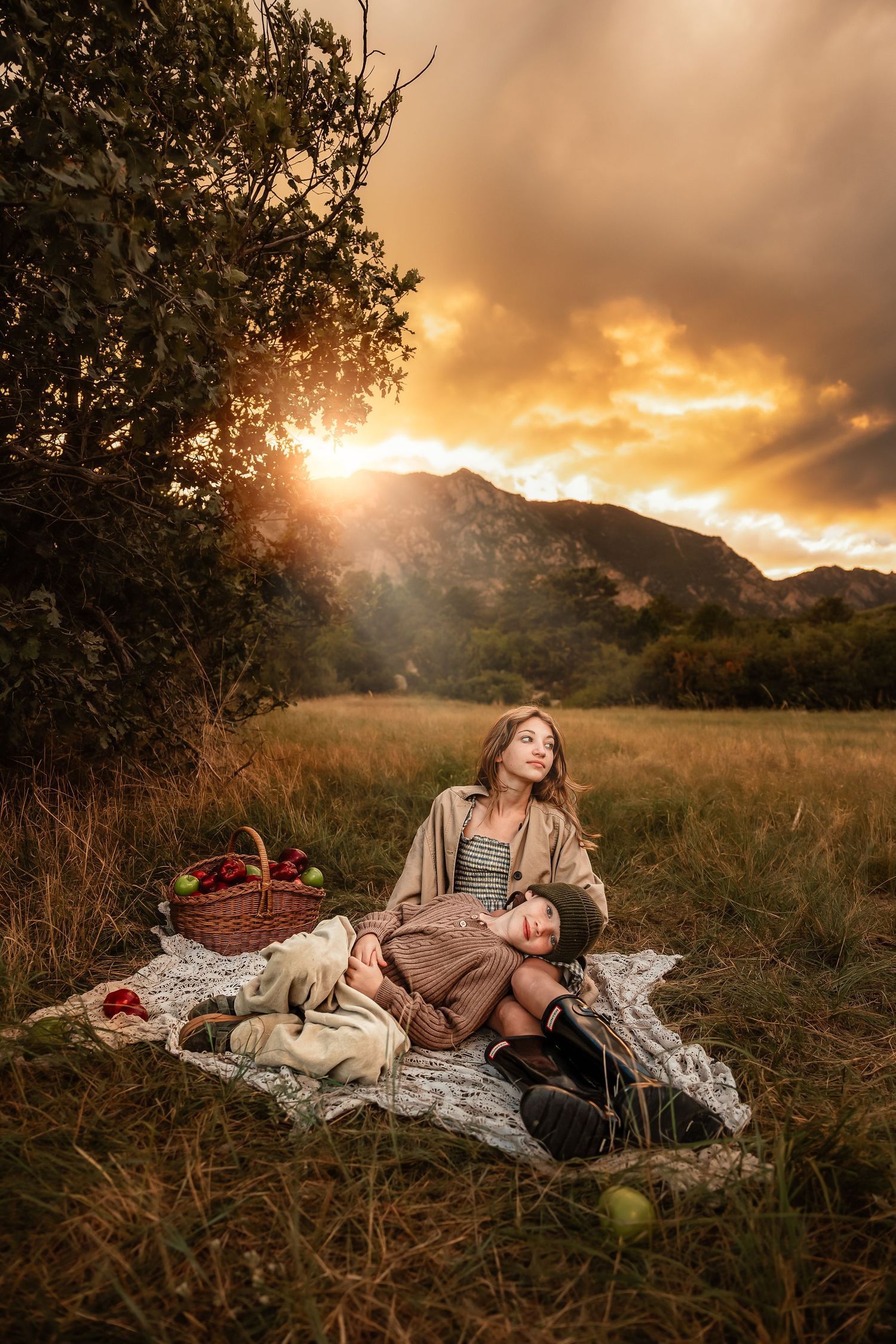 Two women enjoy a picnic on a blanket in a golden field at sunset, with mountains and a fruit basket nearby.