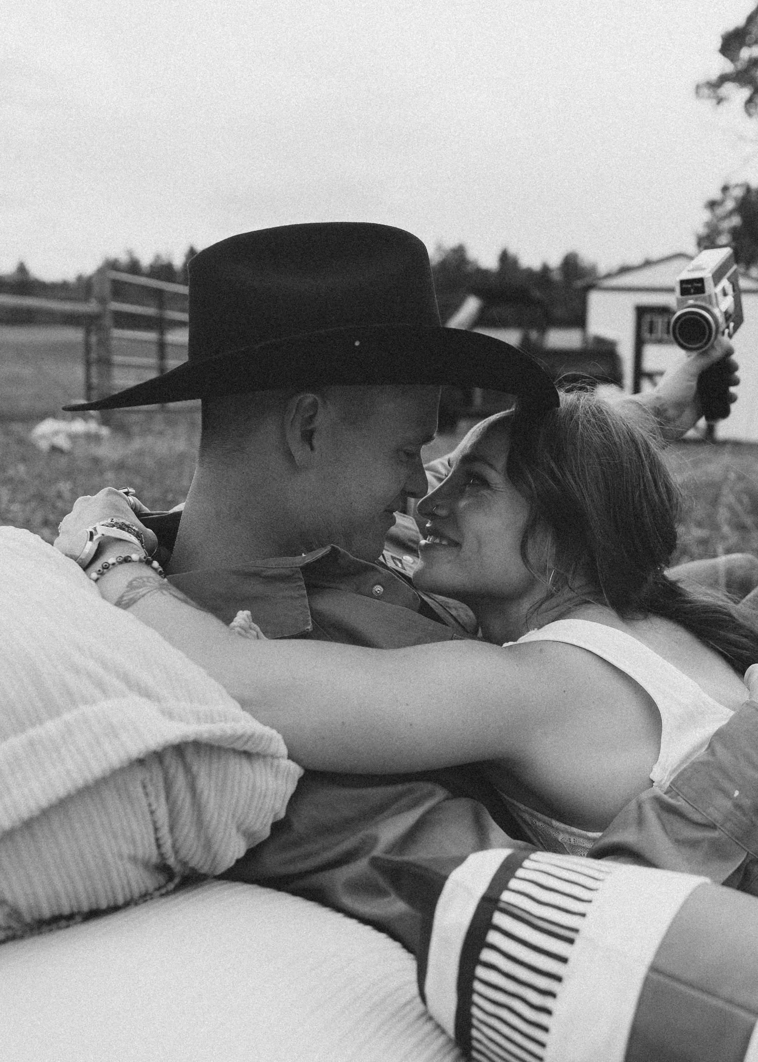 A couple shares a romantic moment outdoors, man in cowboy hat, black and white photo with rustic farm background.