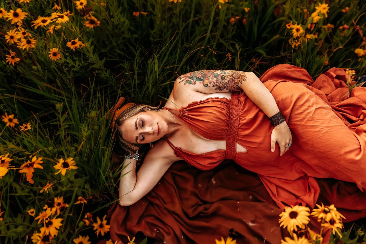 Woman in orange dress lying among yellow wildflowers in a lush green field, showcasing a floral tattoo.