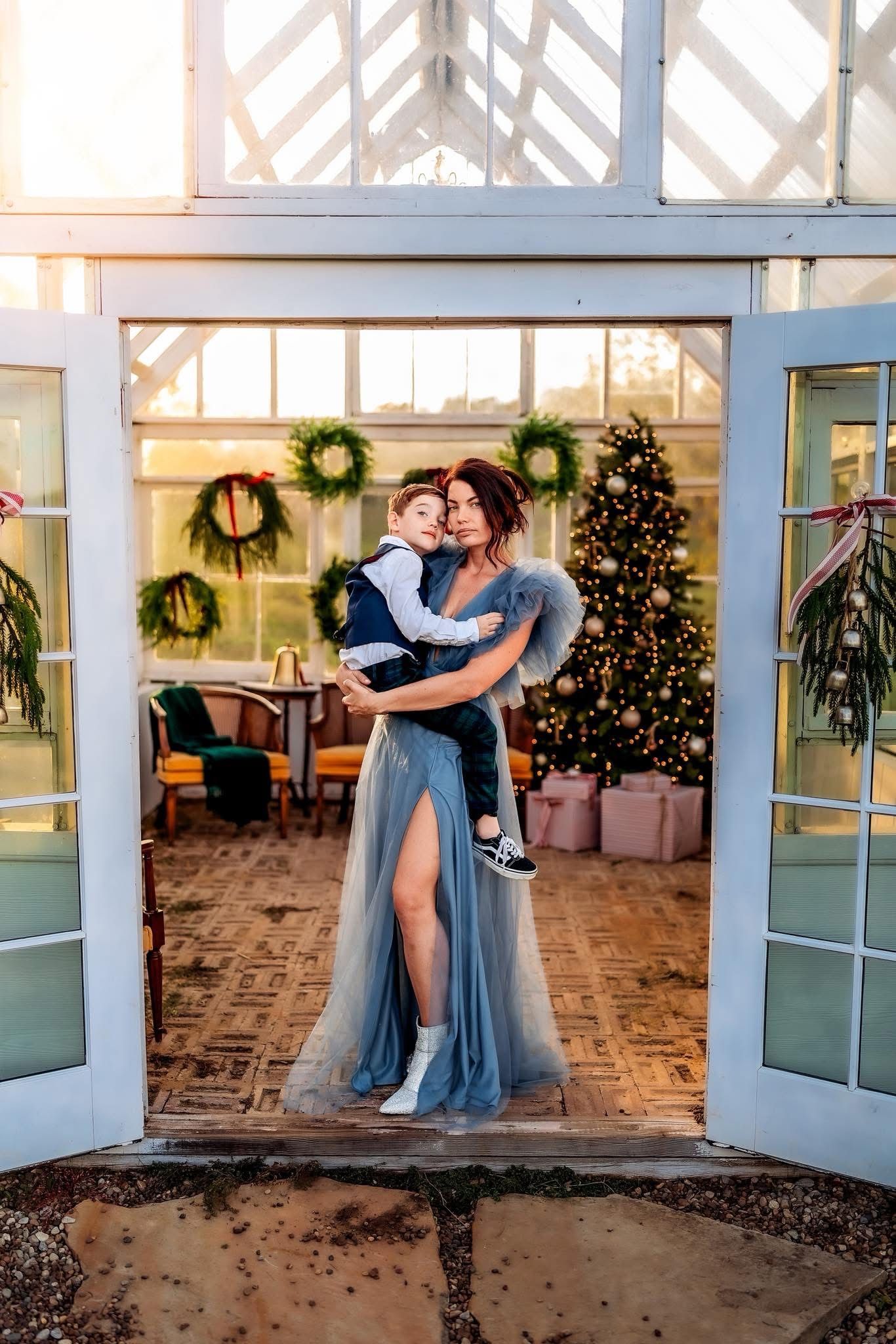Woman in blue tulle gown holding a child in a festive greenhouse doorway with Christmas tree and wreaths.