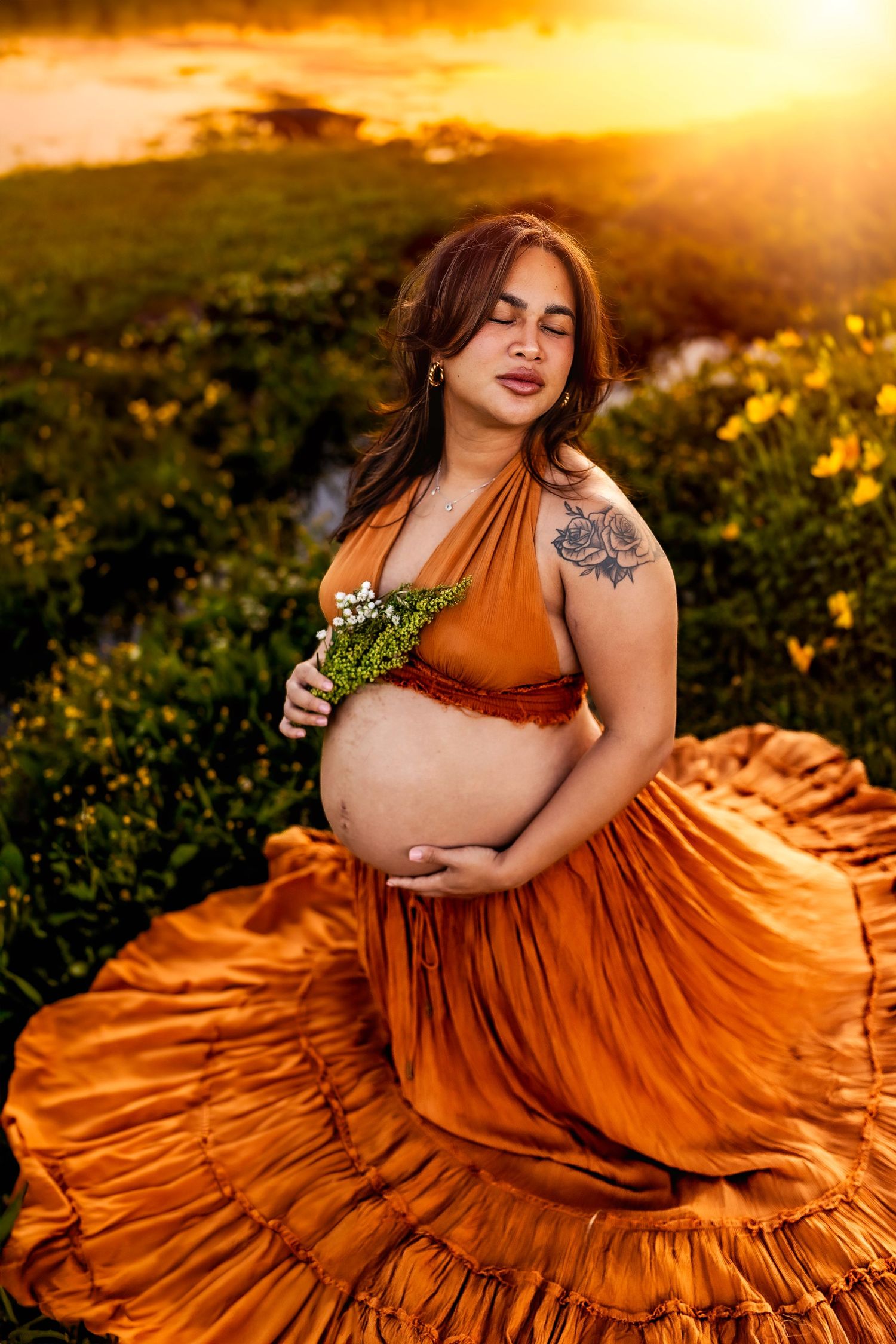 Pregnant woman in orange dress holding flowers at golden sunset in a lush green field.
