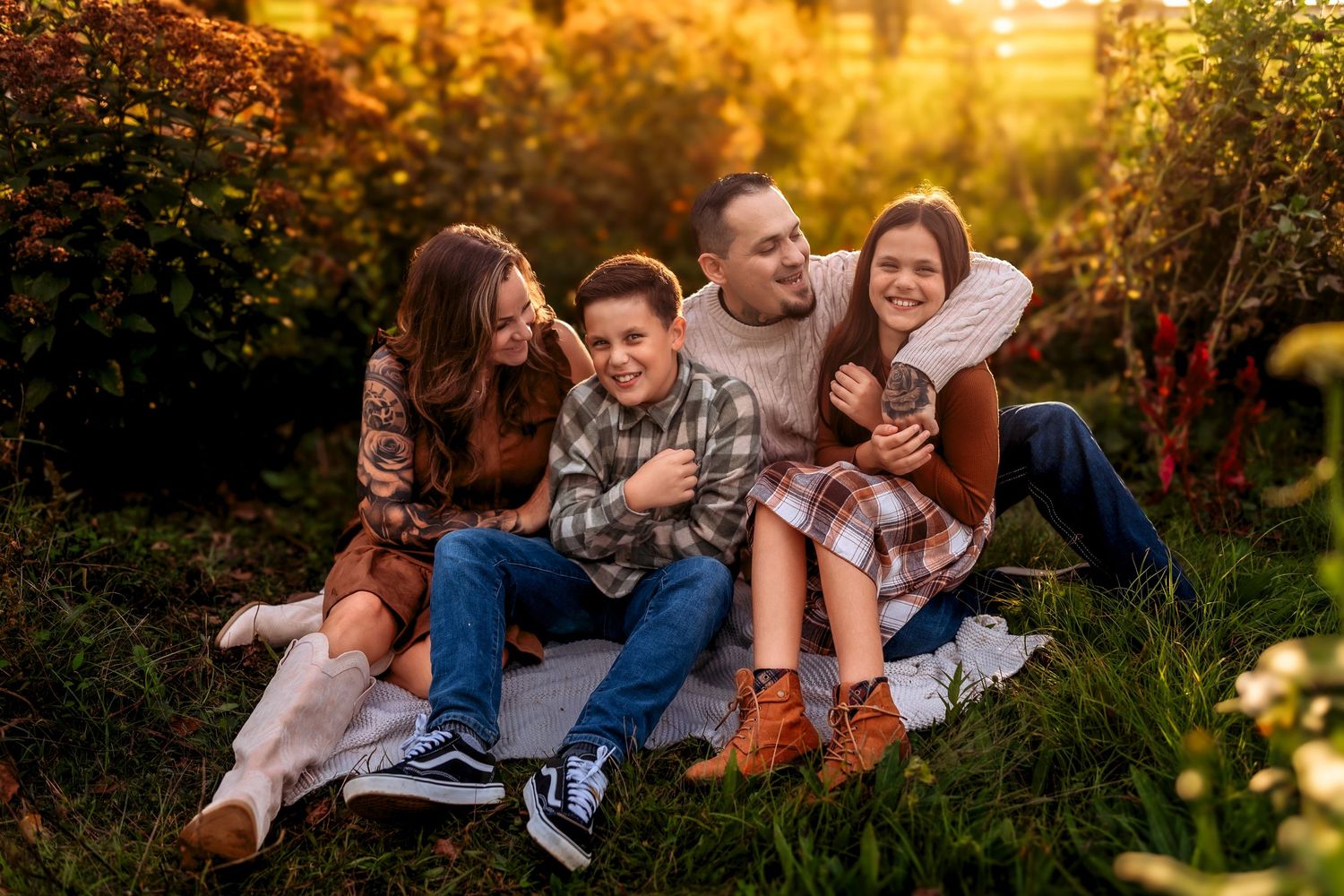 Happy family of four sitting together laughing on blanket in lush green garden at golden hour.