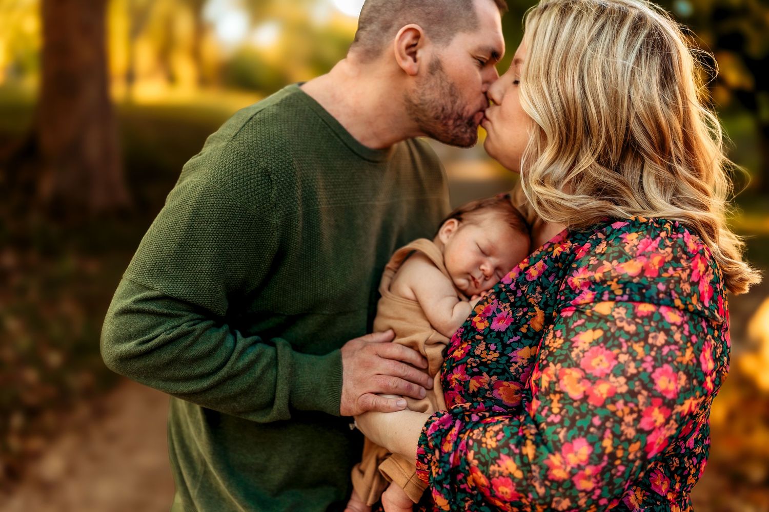 Couple kissing over sleeping newborn baby in colorful floral dress in warm autumn outdoor setting.