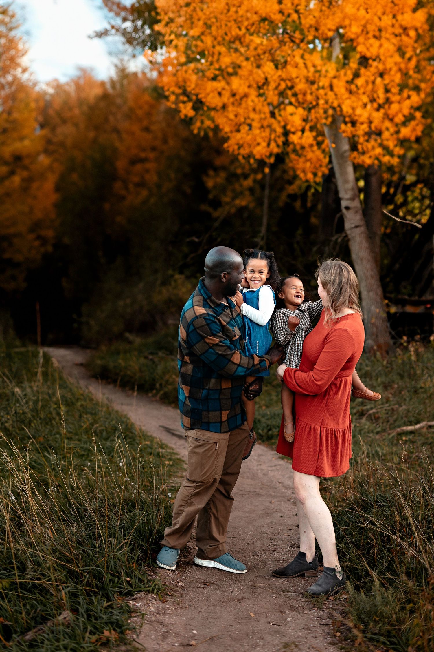 Family of four in a plaid shirt and red dress holding two young children on a dirt path surrounded by golden autumn trees.