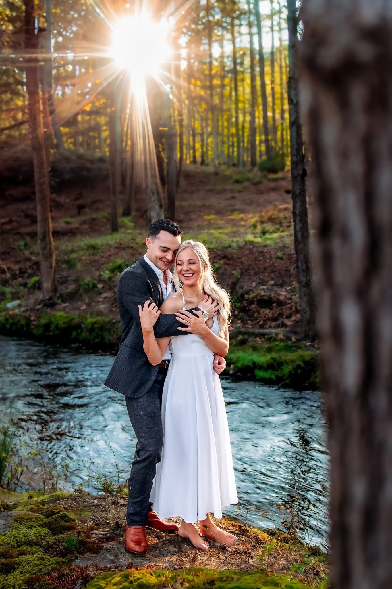 Happy couple embraces by a forest stream with sunlight bursting through autumn trees.
