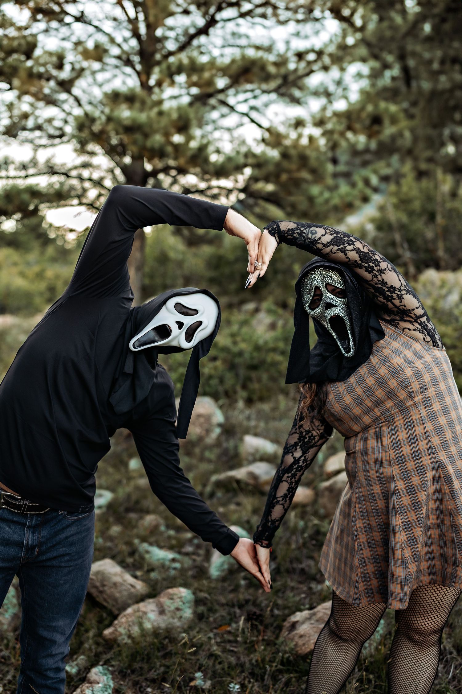 Two people in Ghostface masks join hands to form a heart shape while posing among rocks and pine trees in a forest.