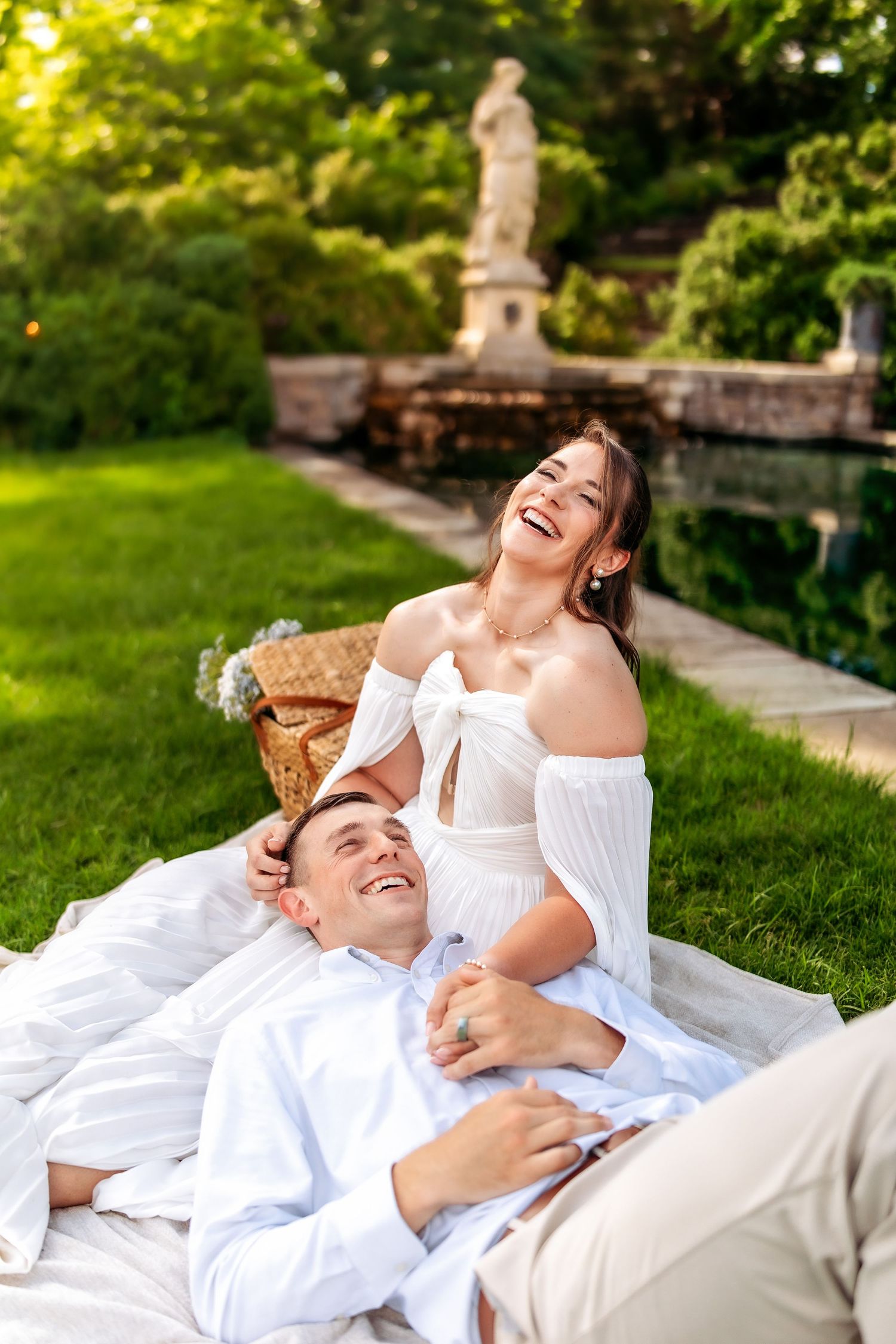 Happy couple in white outfits laughing together on a blanket in a lush garden with a stone statue.