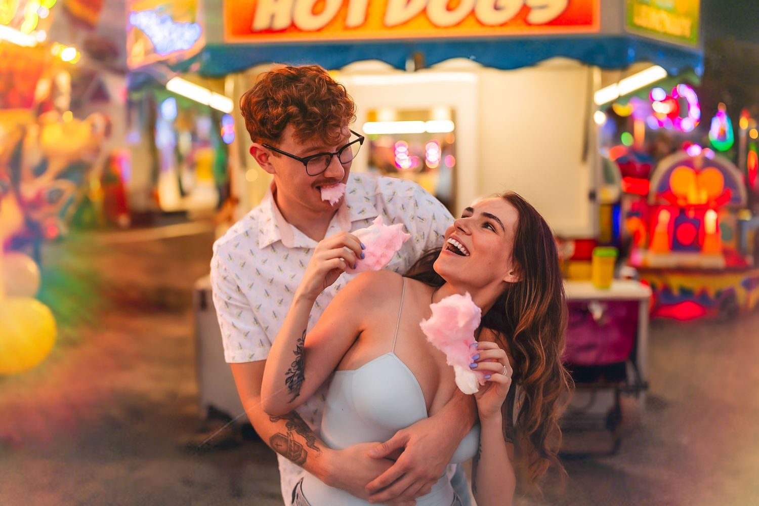 Couple sharing cotton candy at a colorful carnival fair with a hot dog stand in the background at night.