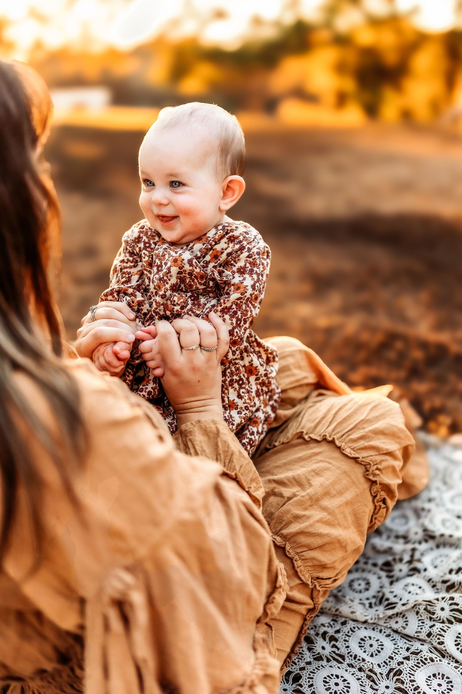 A smiling baby in a floral outfit sits outdoors at golden hour, held by a mother in a mustard dress.