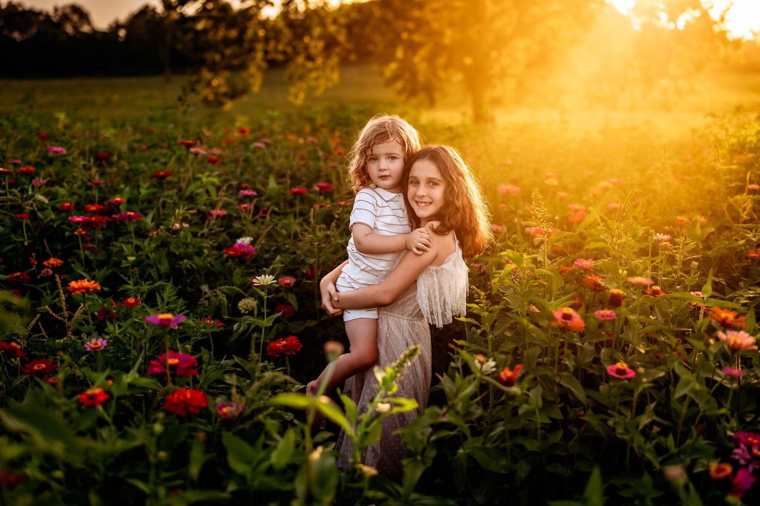 Mother holding toddler in golden sunset light among vibrant red and pink zinnia flowers in a lush garden field.