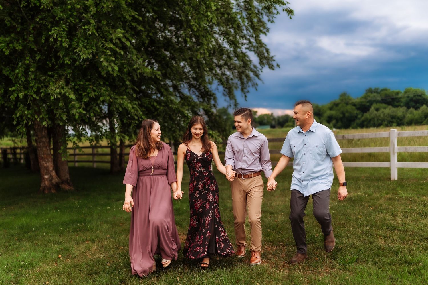 Family of four walking hand-in-hand on green grass near white fence and trees under dramatic cloudy sky.