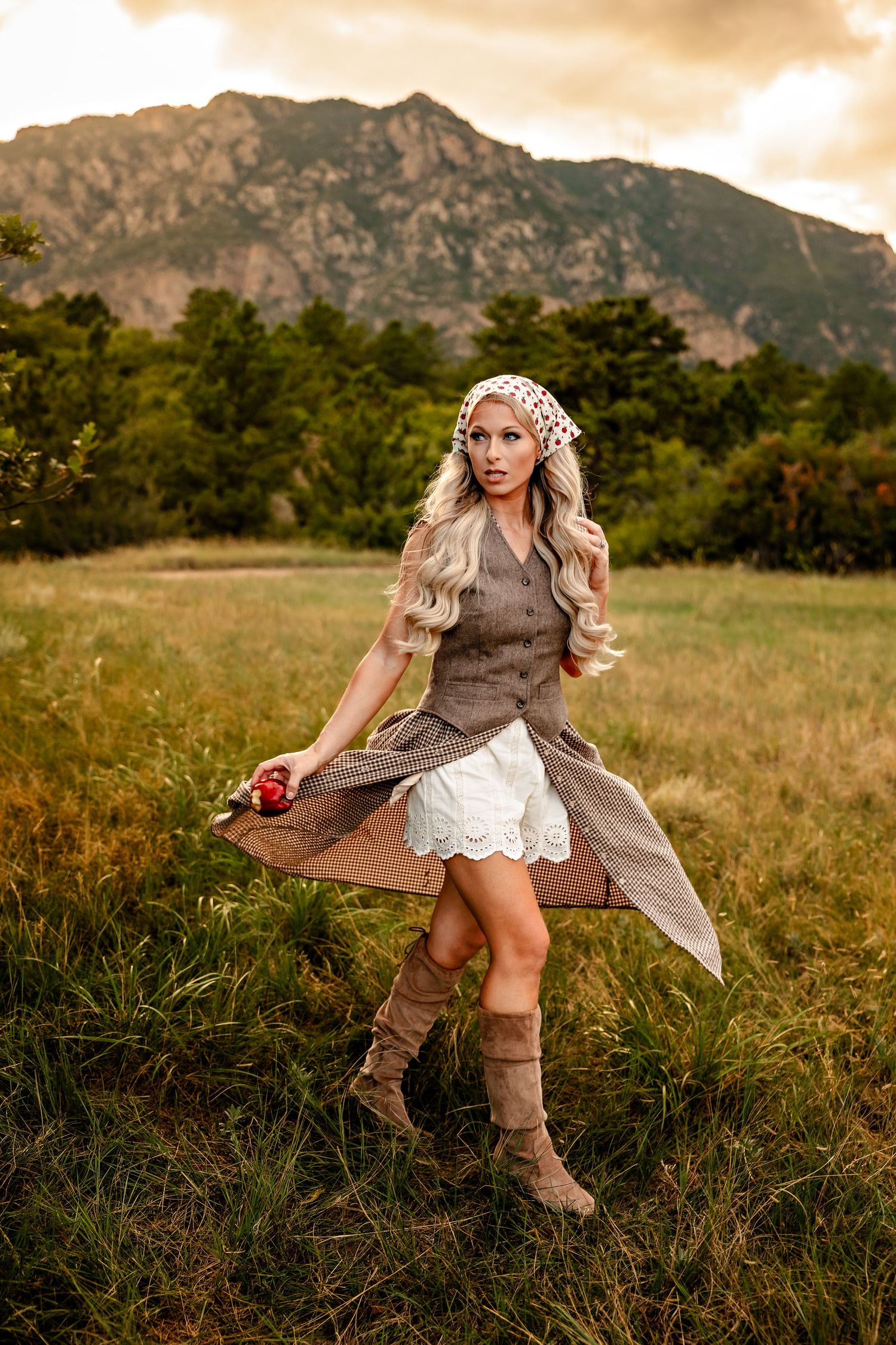 Woman with long blonde hair in a bandana, brown vest and suede boots holding a red apple in a grassy meadow with mountains behind.