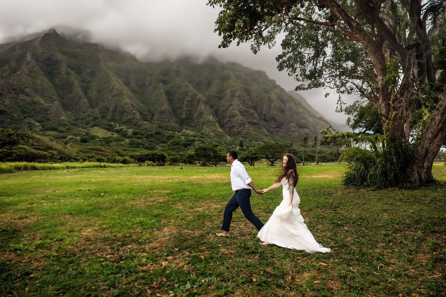 Bride and groom holding hands running through green meadow with misty mountain backdrop in Hawaii.