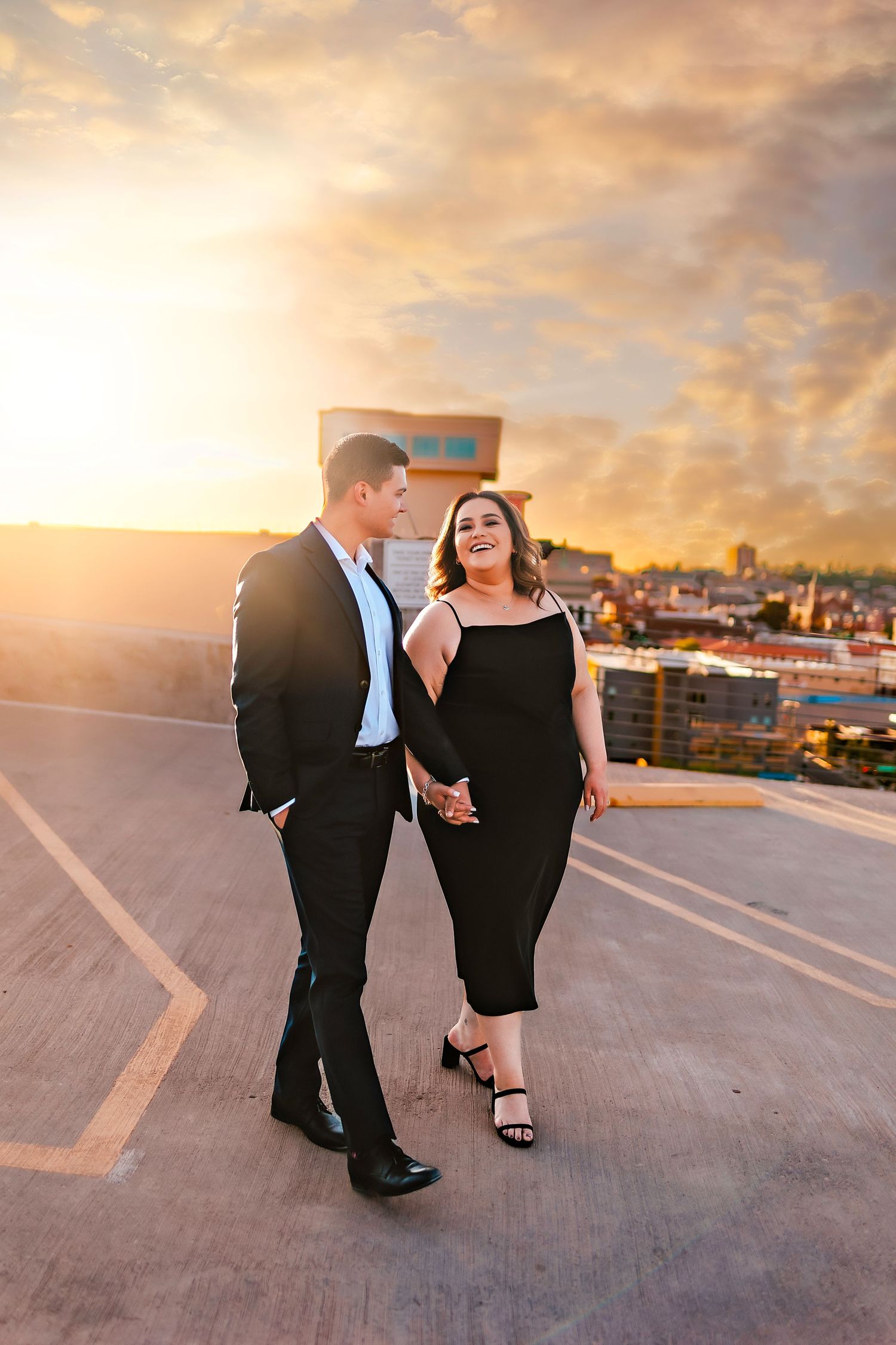 Couple holding hands on rooftop at golden sunset, man in black suit, woman in black dress, city skyline behind them.