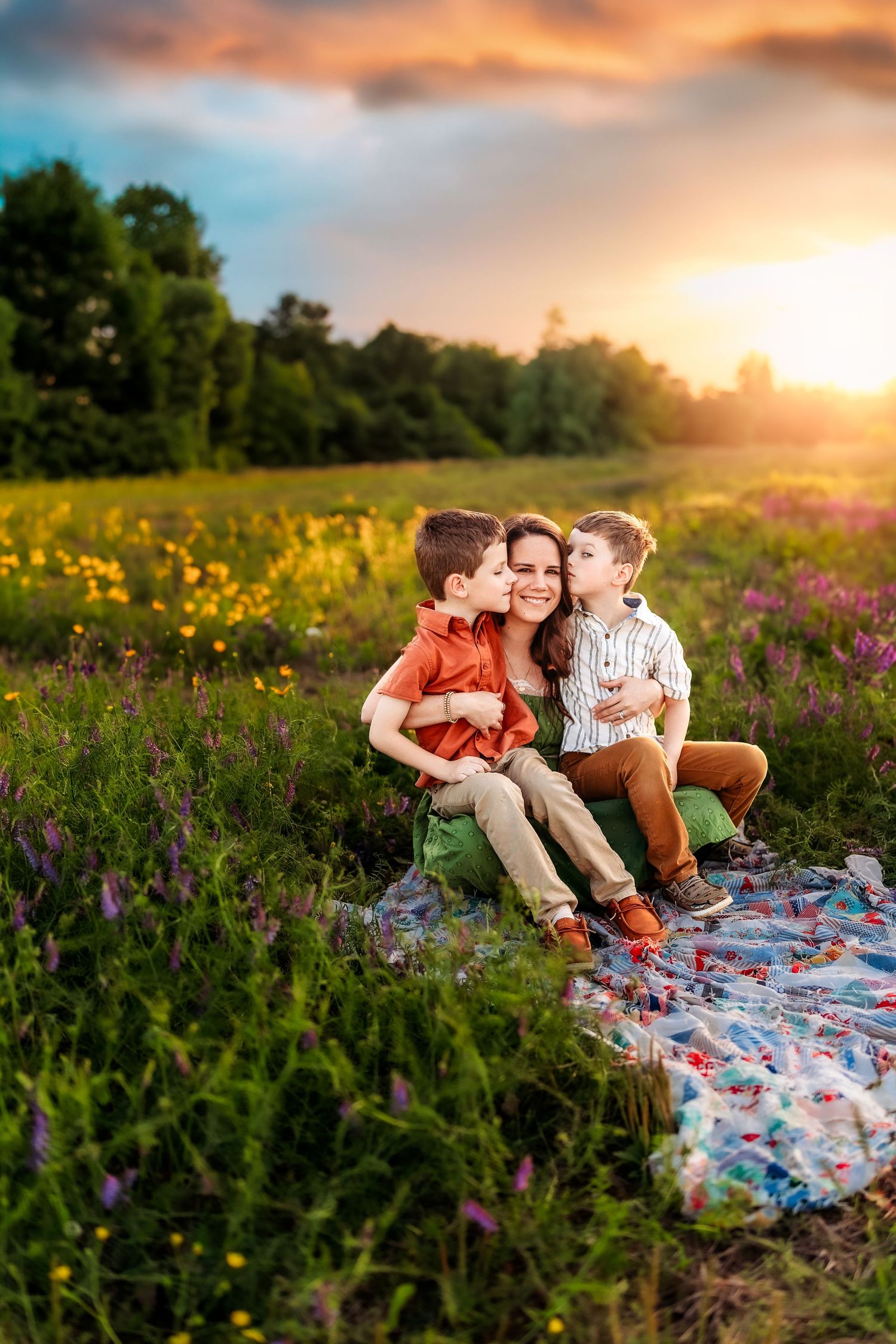 A mother sits in a flower field at sunset, smiling as her two young sons kiss her cheeks on a blanket.