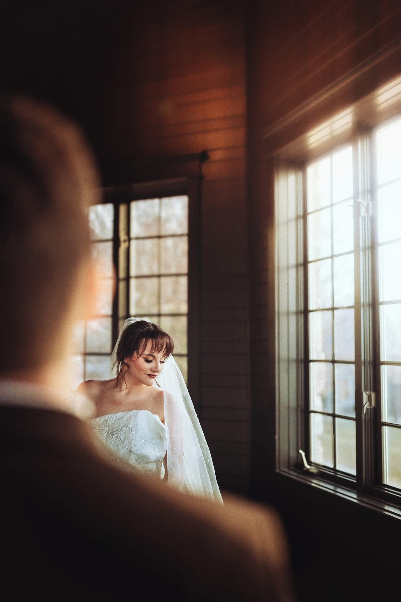 Bride in white gown and veil stands by sunlit windows in a rustic wood-paneled room during wedding ceremony.