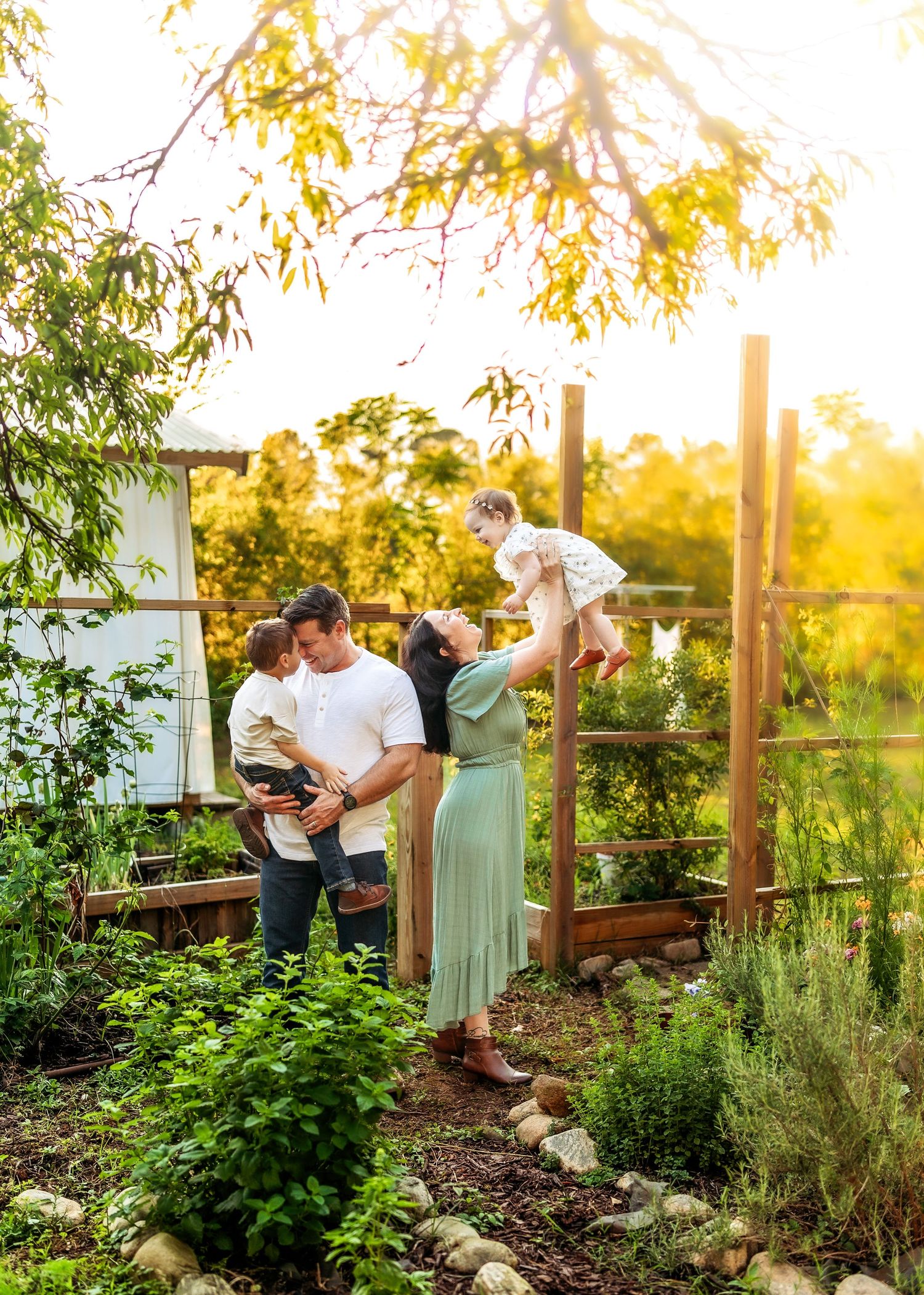 A family enjoys a golden hour moment in a lush garden, parents lifting two young children near a wooden fence.