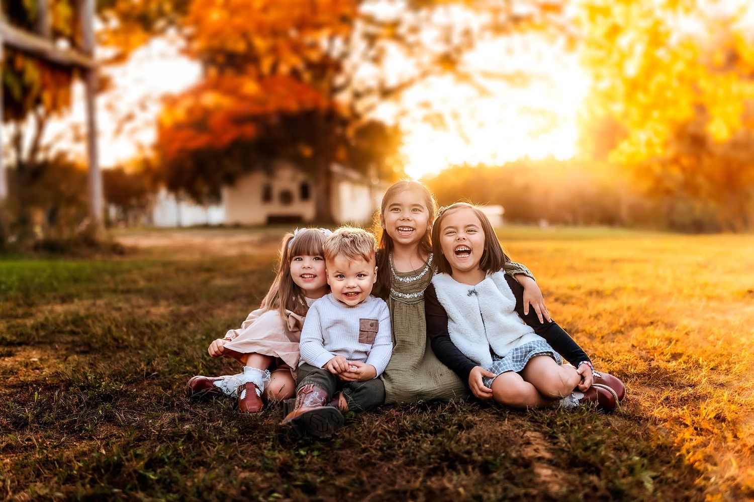 Four young children sitting together on grass smiling, surrounded by vibrant autumn foliage in warm golden sunlight.