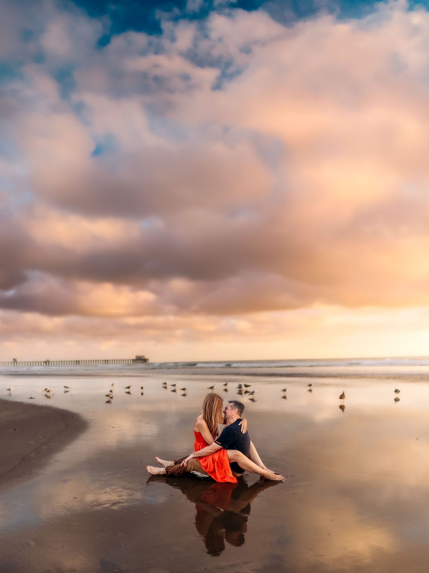Couple sitting on wet beach sand at sunset with colorful clouds, ocean pier, and seagulls in background.