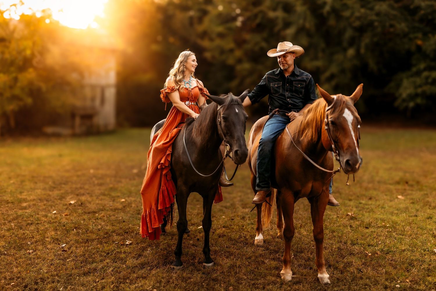 A couple in western attire rides horses at golden sunset, woman in red gown, man in cowboy hat, on lush green field.