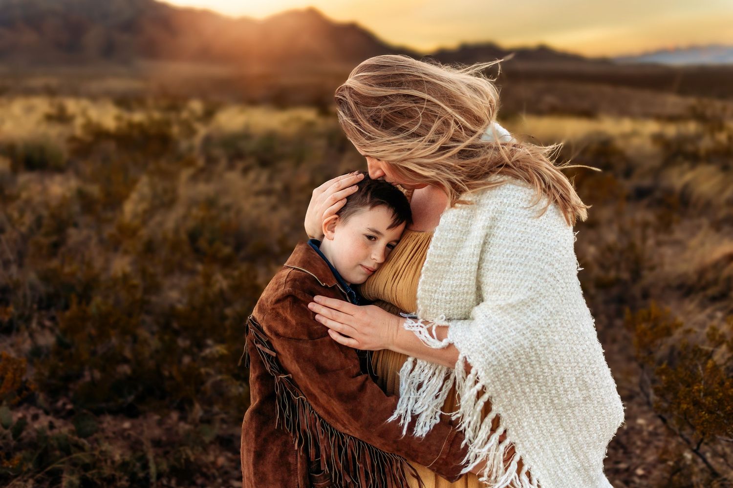 A mother tenderly embraces her young son in a sunlit desert field, both wearing warm fringed wraps at golden hour.