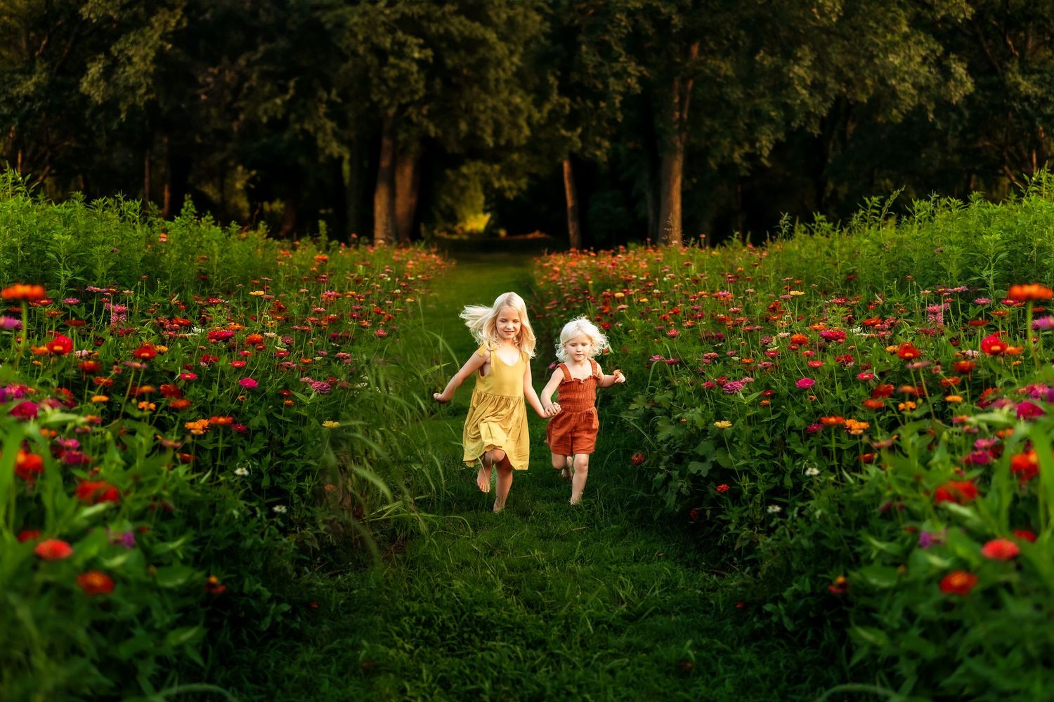Two young girls in fairy costumes run joyfully through a vibrant flower garden path lined with red blooms.