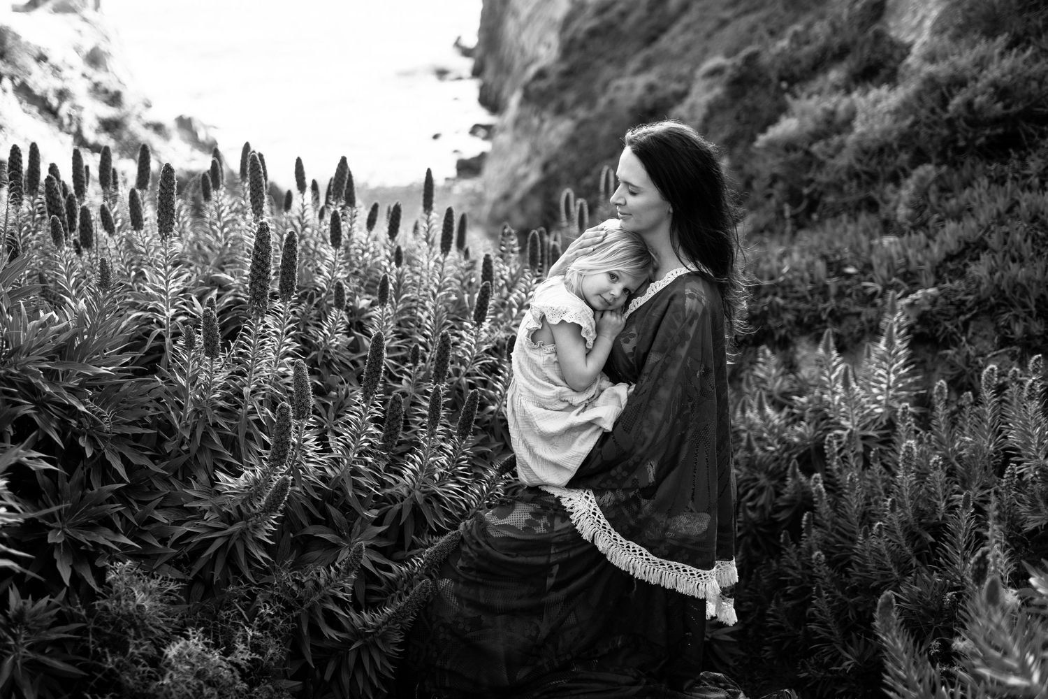 Black and white photo of a woman cradling a baby among tall flowering plants on a rocky hillside.