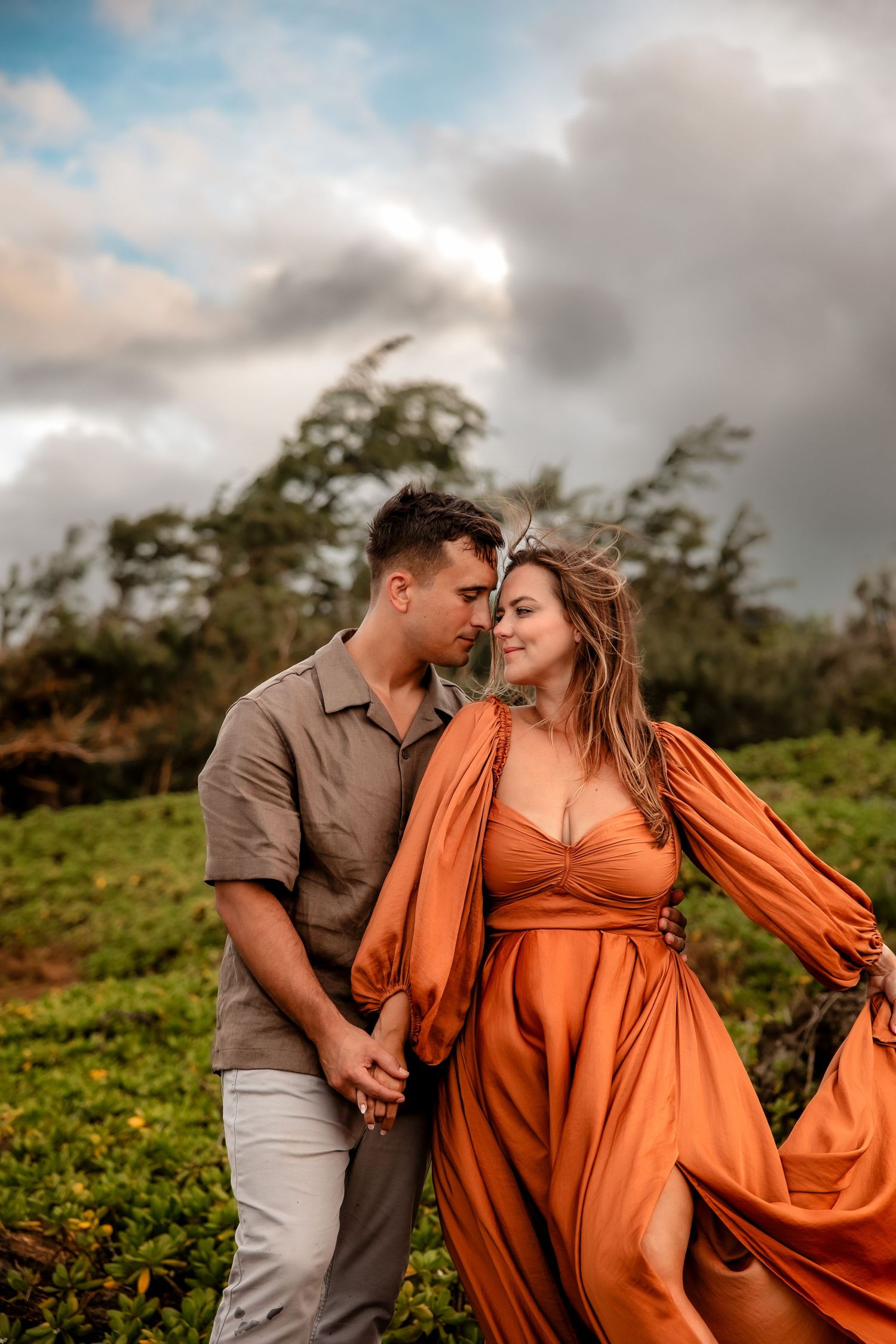 Couple in orange dress and gray shirt holding hands in a lush green field under dramatic cloudy skies.