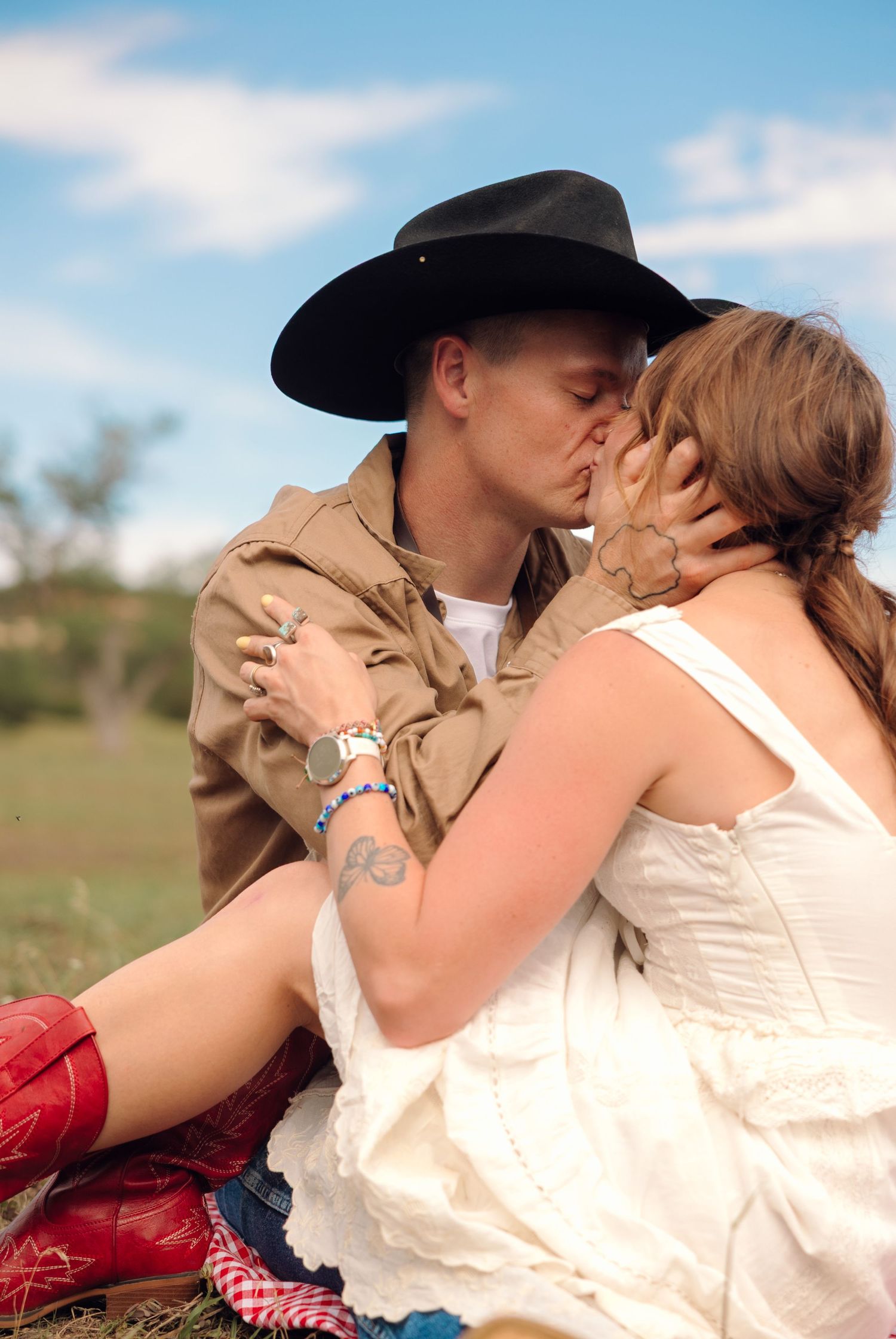A couple in western attire sharing a romantic kiss outdoors, woman in white dress with red boots, man in black cowboy hat.
