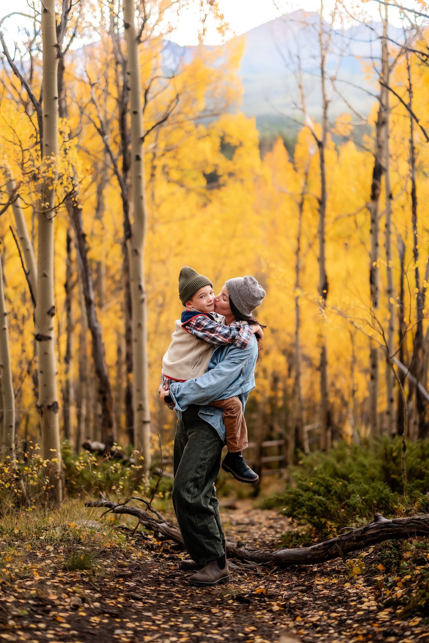 A parent holds a child in a vibrant autumn aspen forest with golden yellow trees lining a scenic mountain trail.
