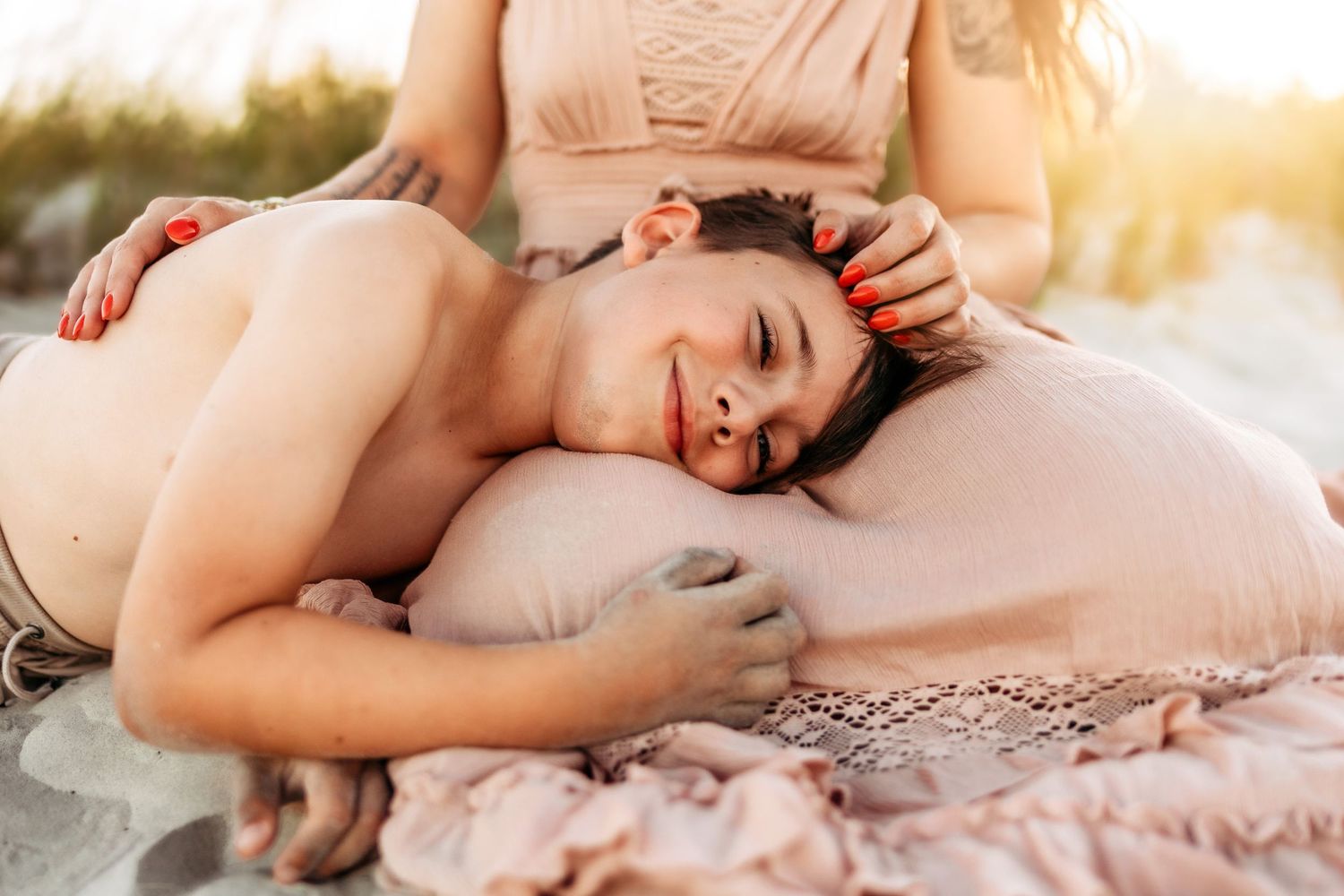 Two women relaxing on a beach blanket at sunset, one resting her head on the other's lap.