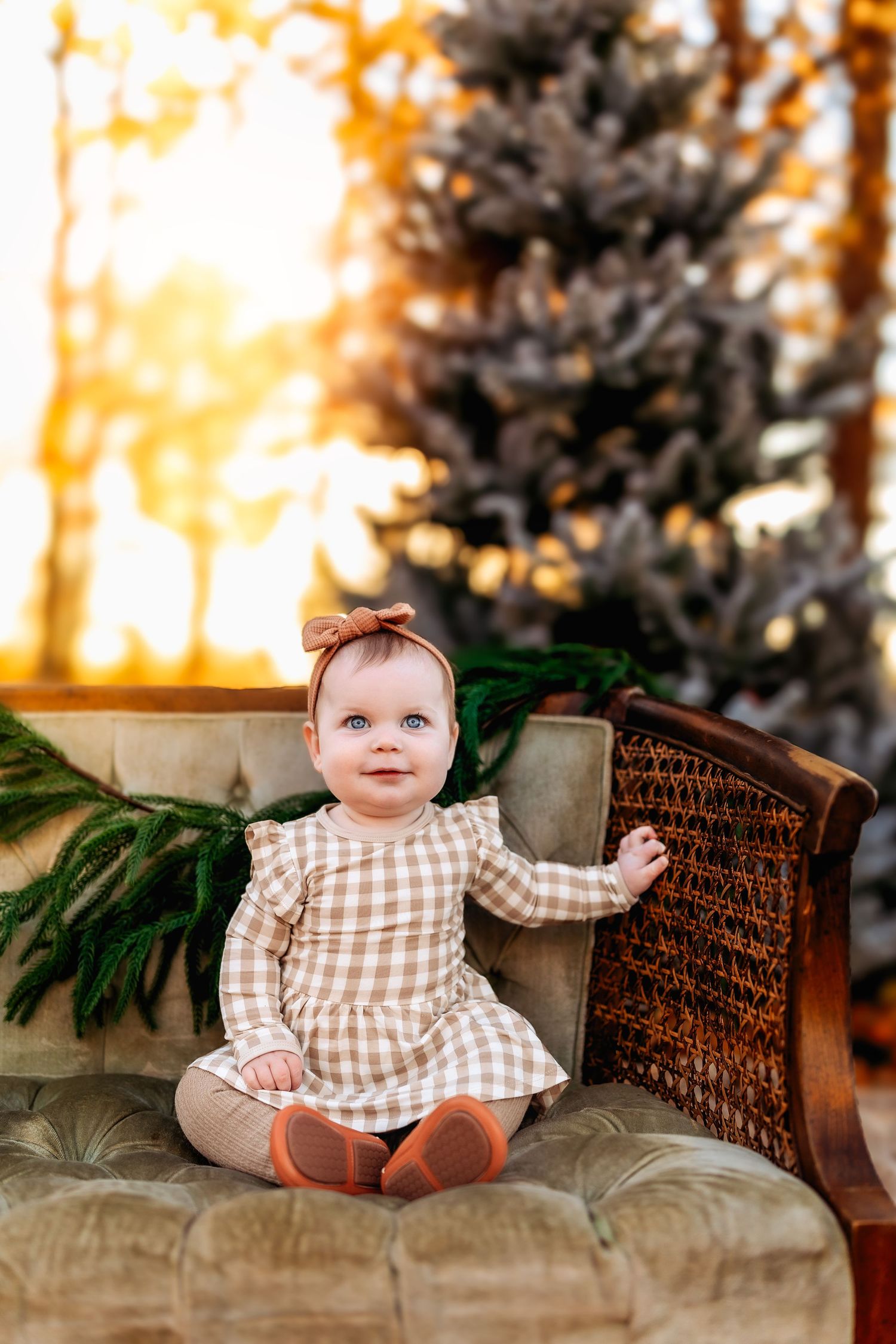 A baby girl in a plaid dress sits on a velvet chair near a Christmas tree in warm golden light.