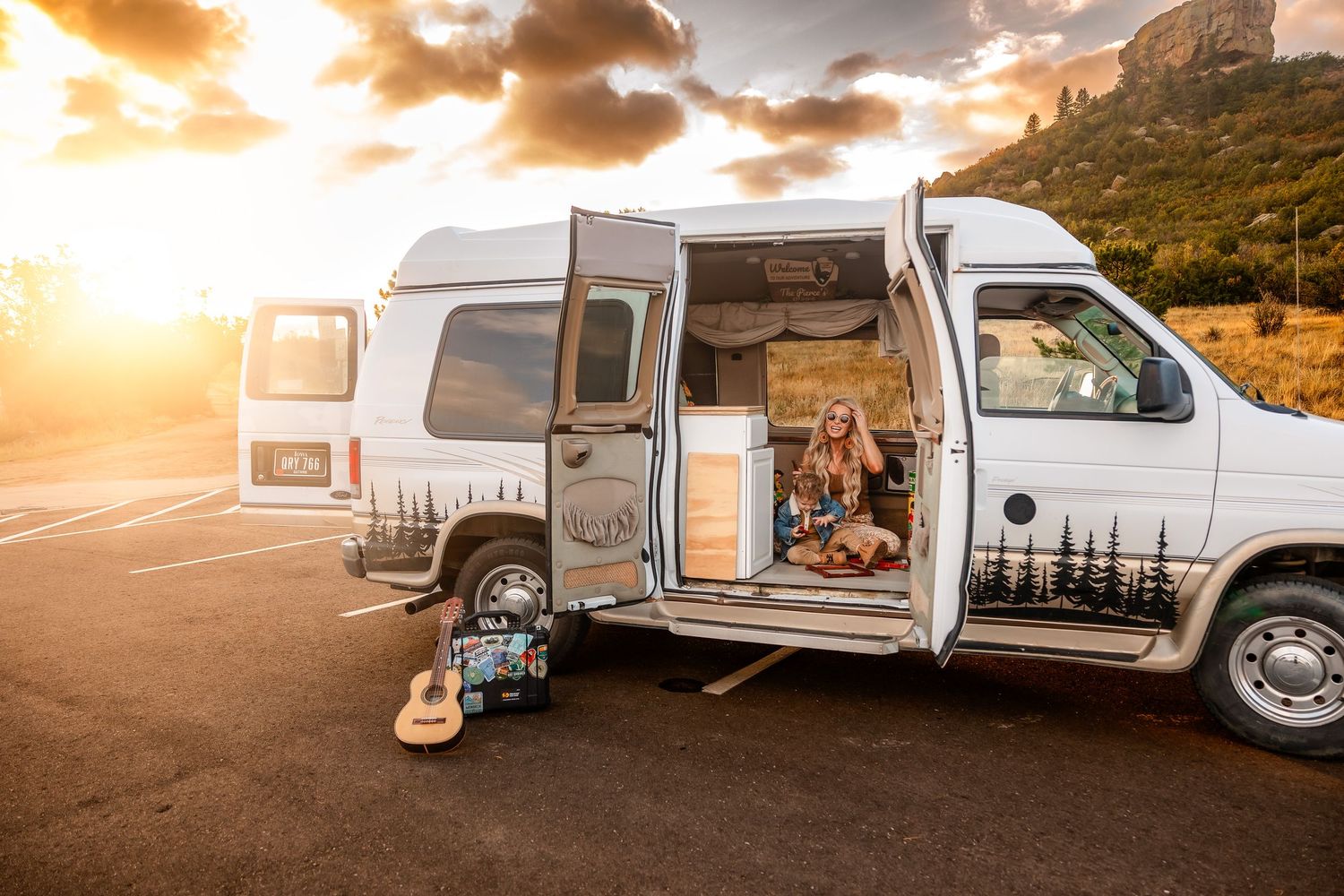 Woman sitting in open converted camper van with guitar at sunset, mountains in background.