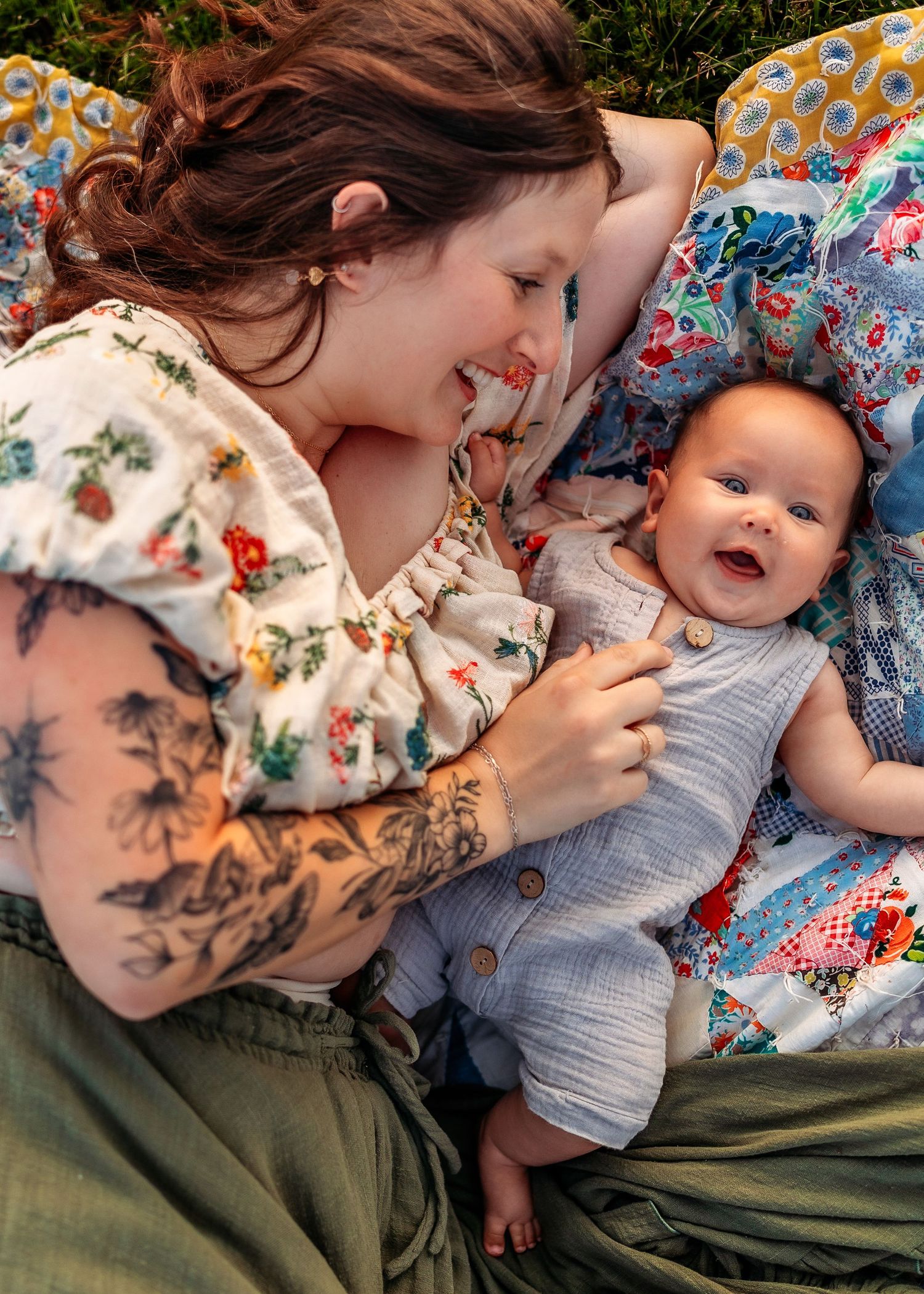 Mother with tattoos smiles at her happy baby lying on a colorful quilt outdoors.
