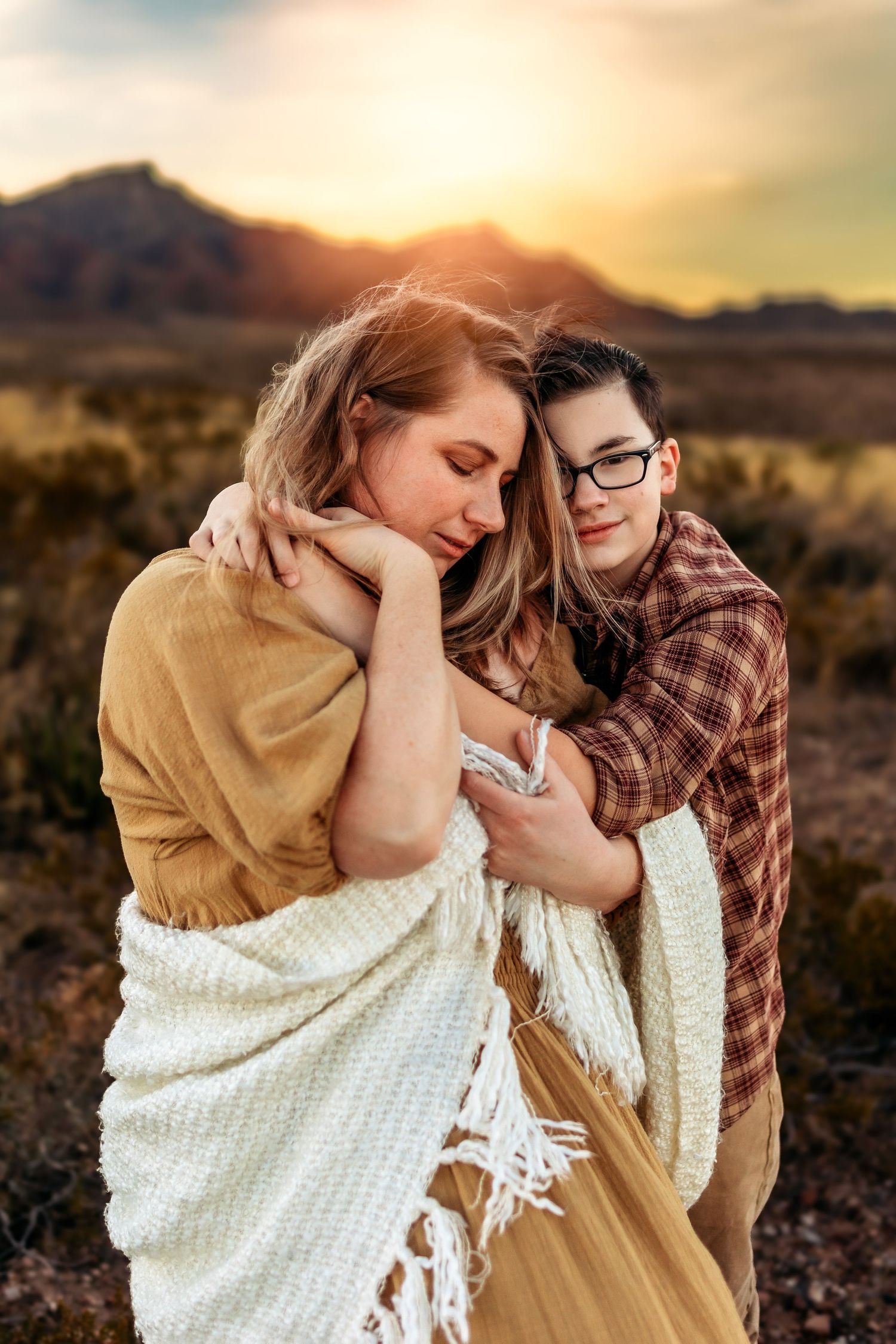 Couple embracing at sunset in a desert landscape, woman in yellow dress with white blanket, man in plaid shirt with glasses.