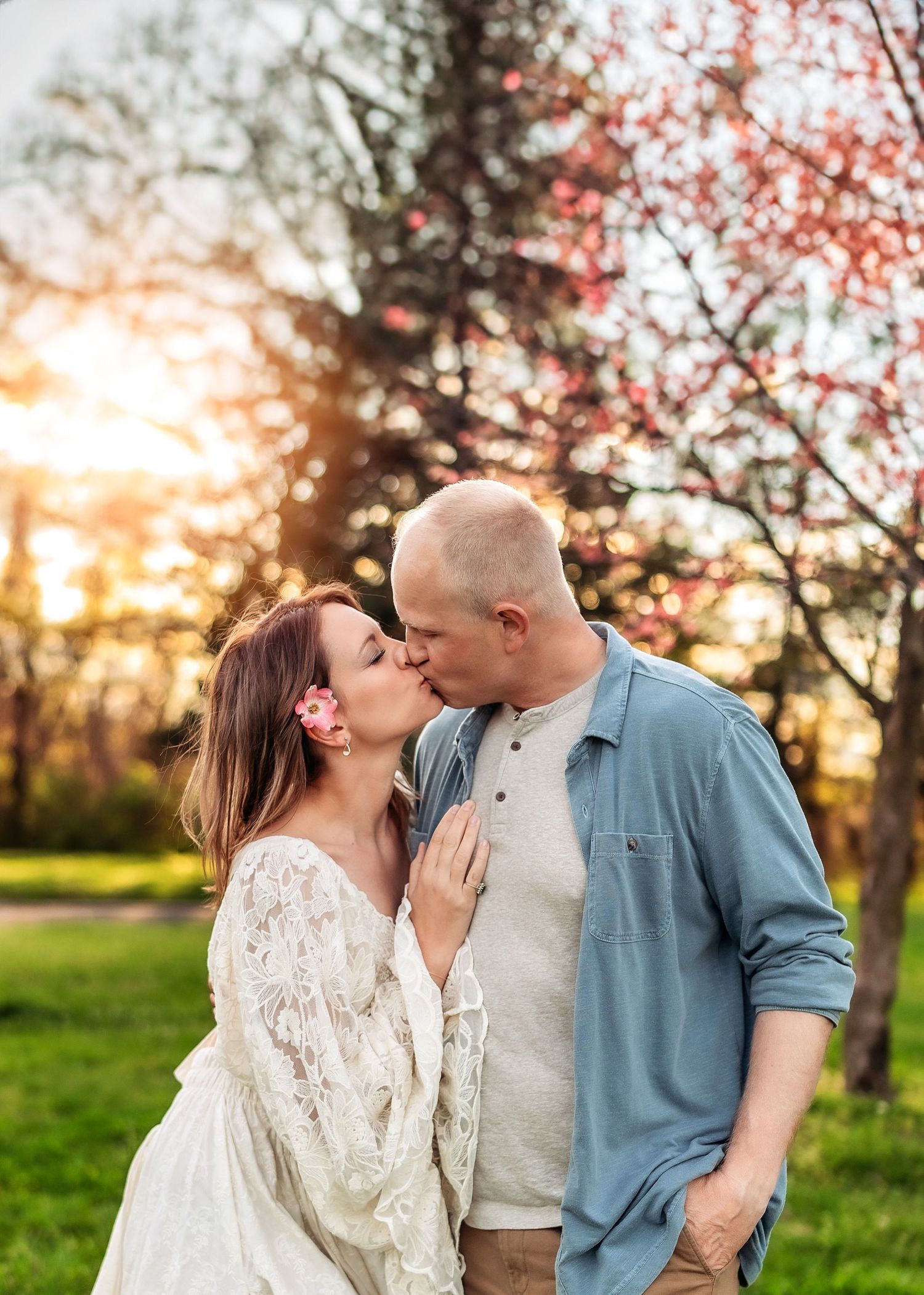 Couple sharing a romantic kiss outdoors at sunset with blooming spring trees in a lush green park.