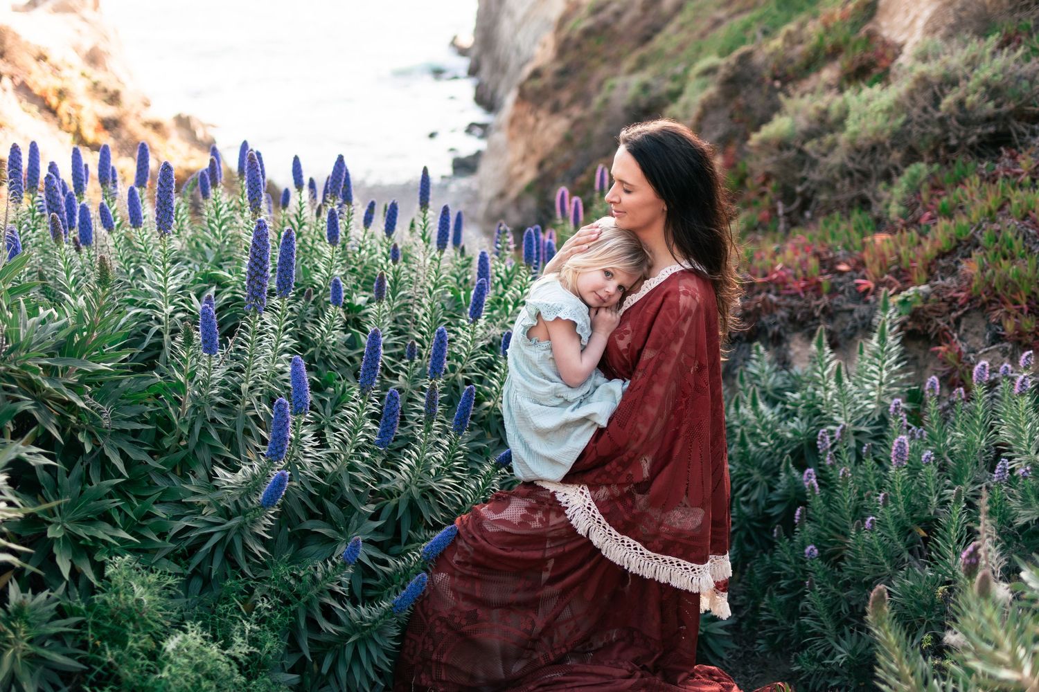 Mother in red dress holds child among blue wildflowers on coastal cliffside.