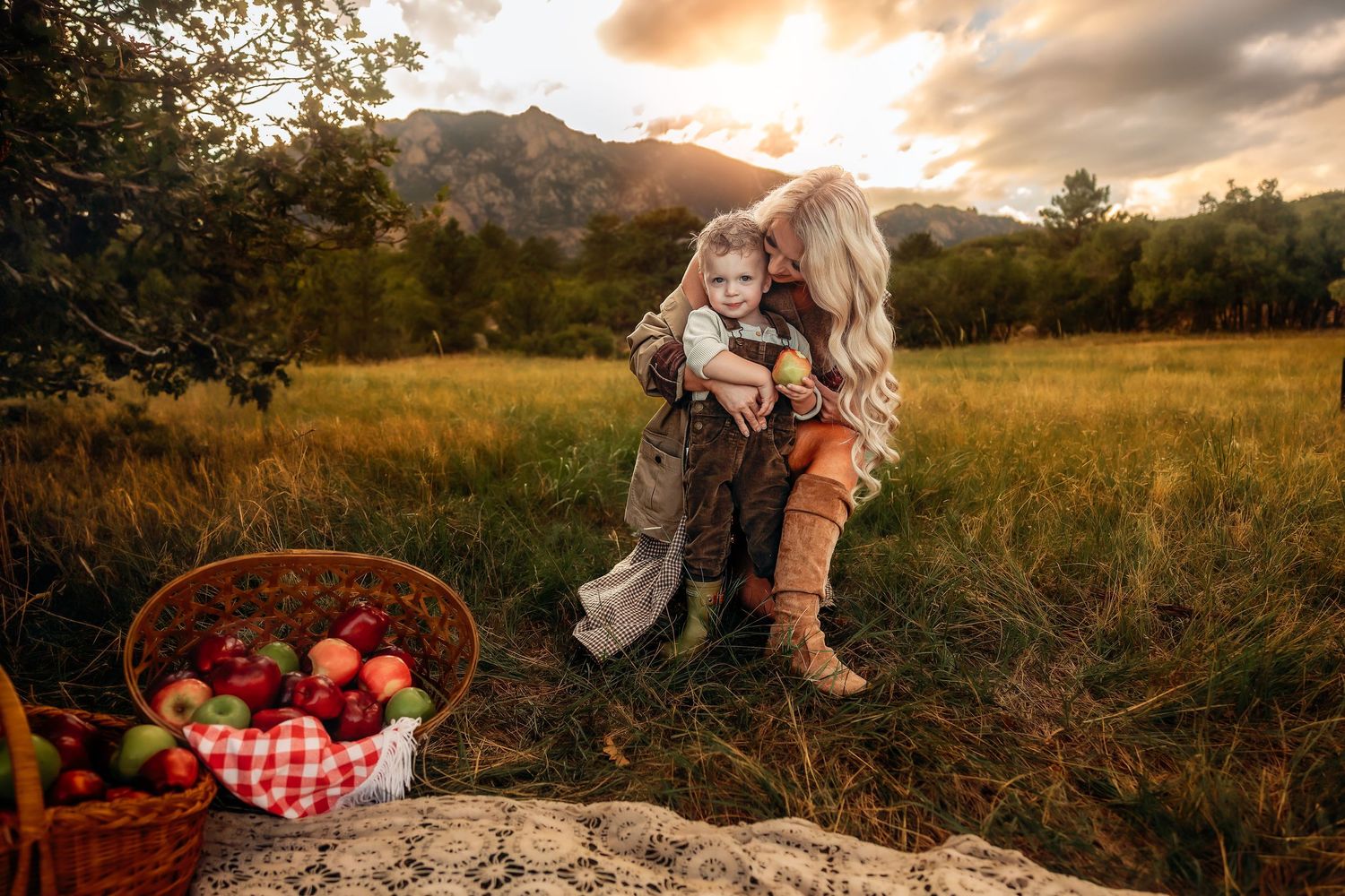 Mother holding her toddler in a golden grassy field at sunset, with a wicker basket of fresh apples and mountains in the background.