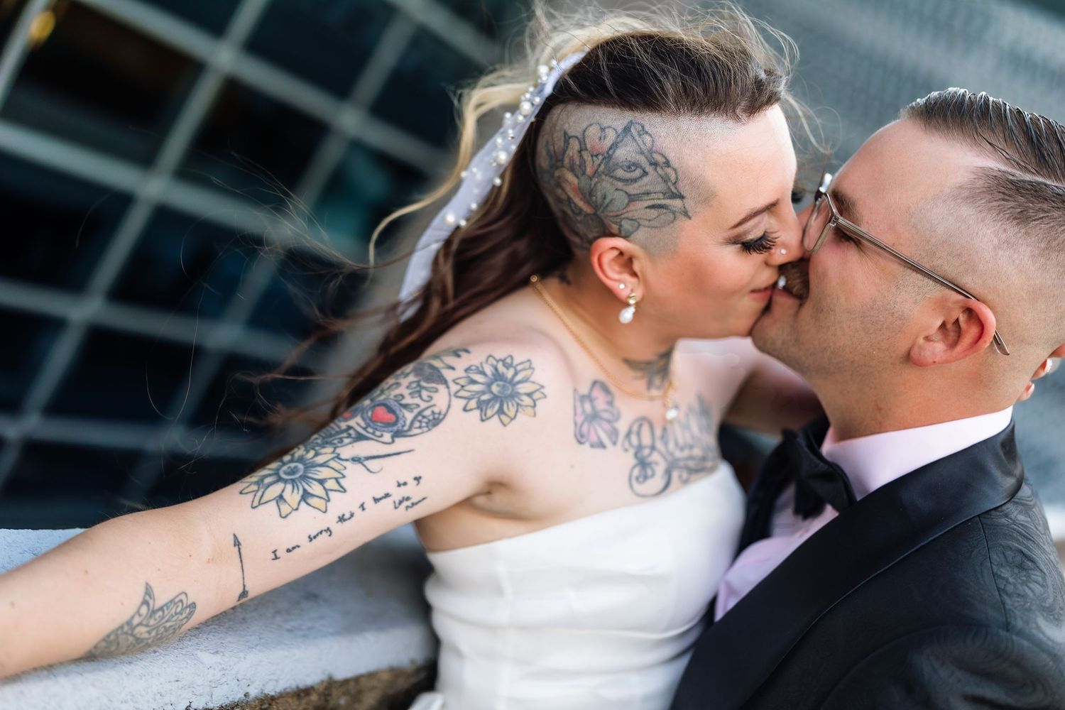 Tattooed bride and groom sharing a kiss on their wedding day, bride wearing white gown with extensive floral tattoos.
