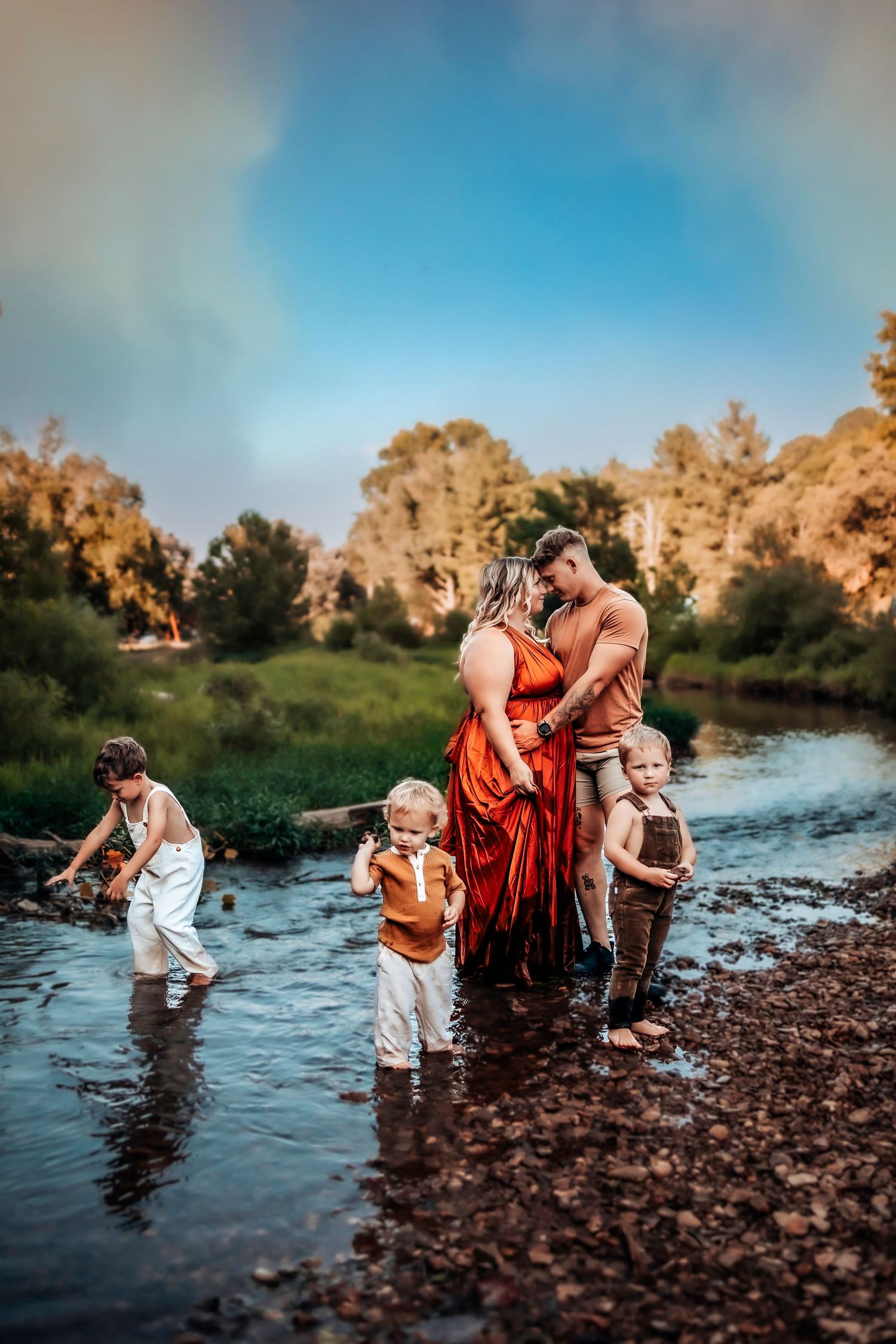 Pregnant couple embracing in a creek surrounded by three young children during a golden hour family maternity photo session.