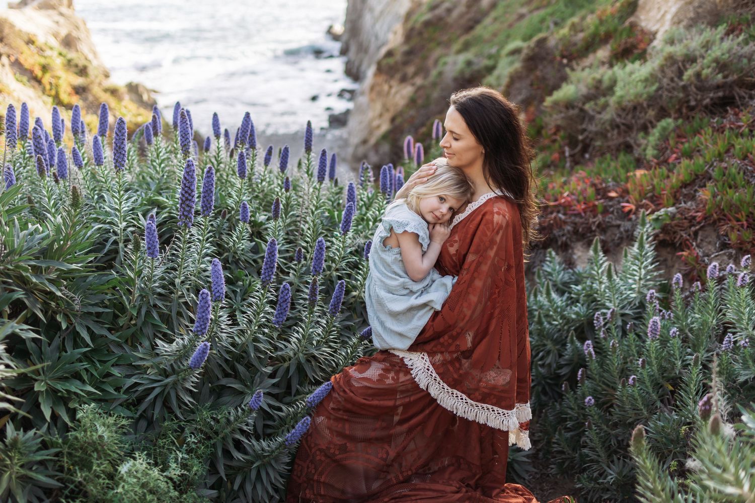 Woman in red dress holding a baby among purple wildflowers on a coastal cliffside.
