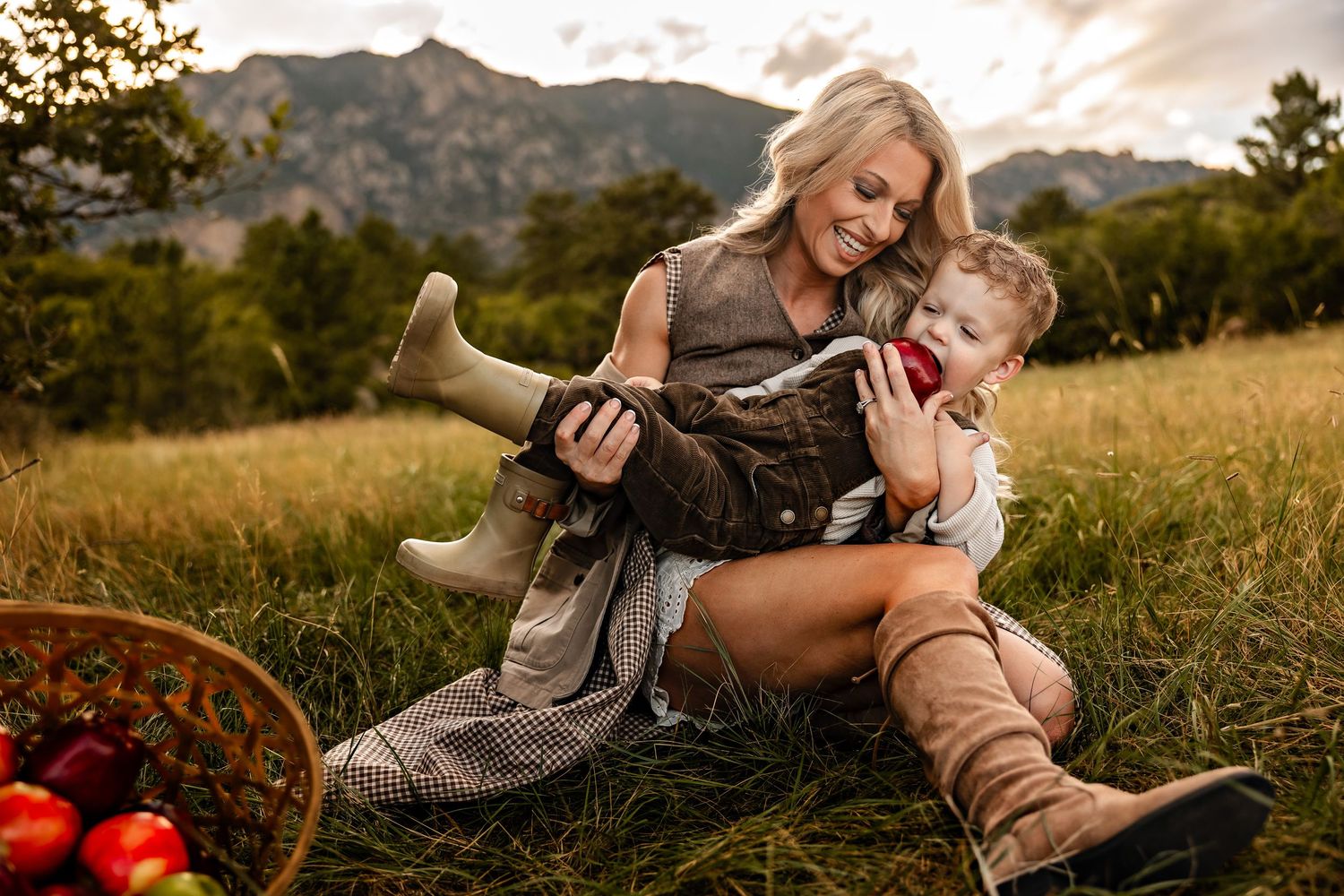 Smiling blonde mother in boots playfully holds her laughing toddler in a sunny grassy field with a basket of red apples and mountains behind