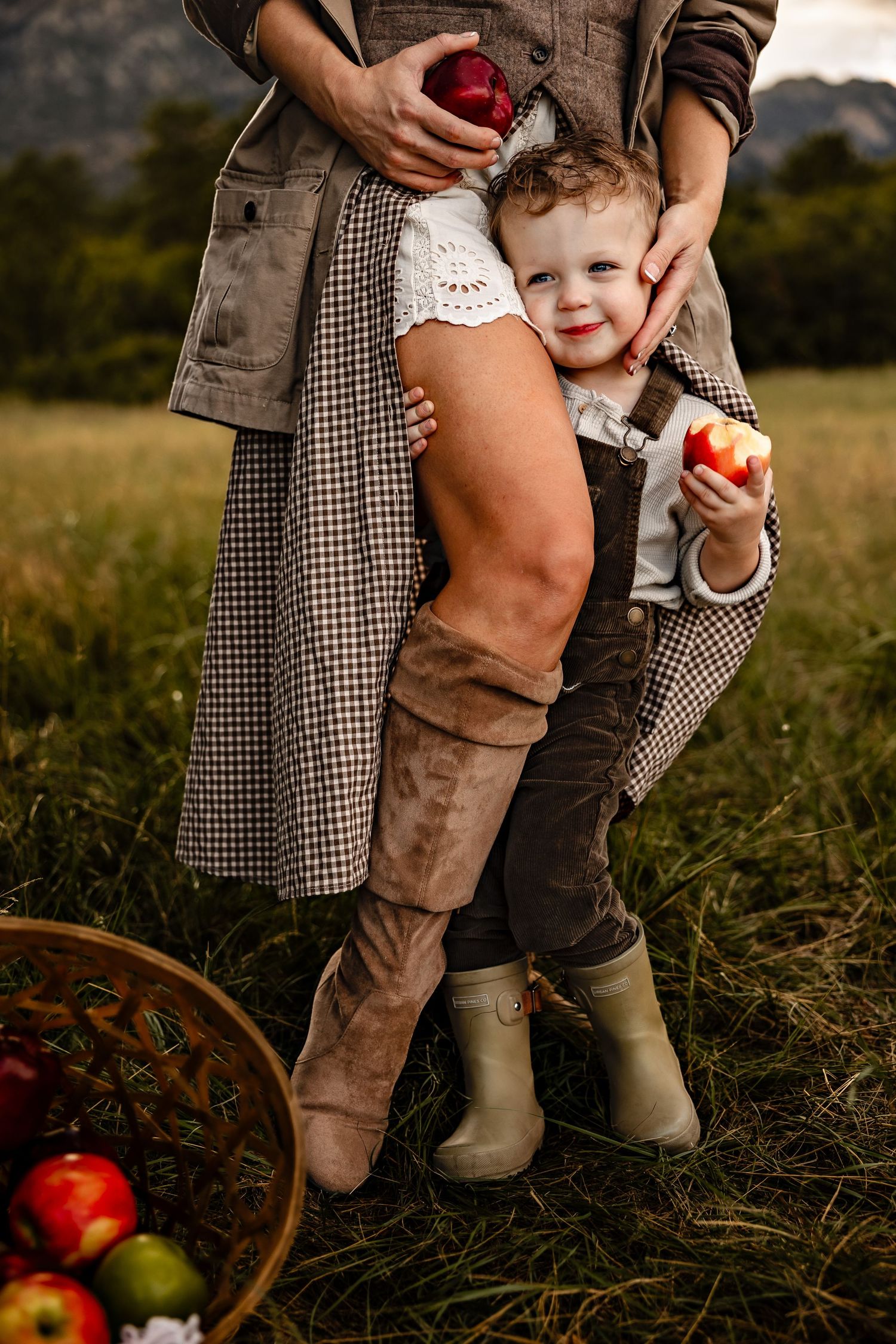Smiling toddler in corduroy overalls and rain boots holds a bitten apple beside a woman in tall suede boots in a grassy apple orchard.
