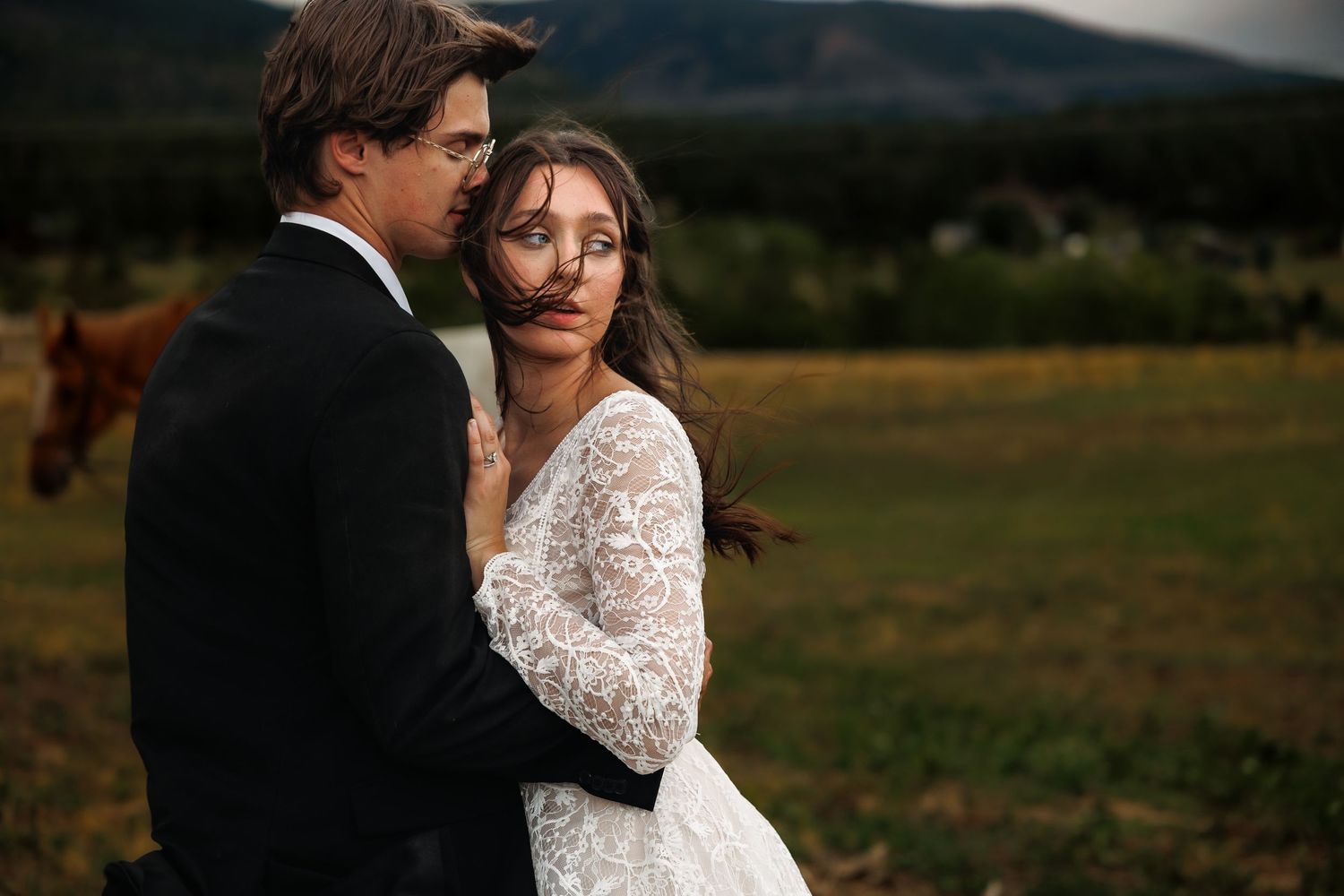Bride in lace long-sleeve gown embraces groom in black suit outdoors with mountains and horses in background.