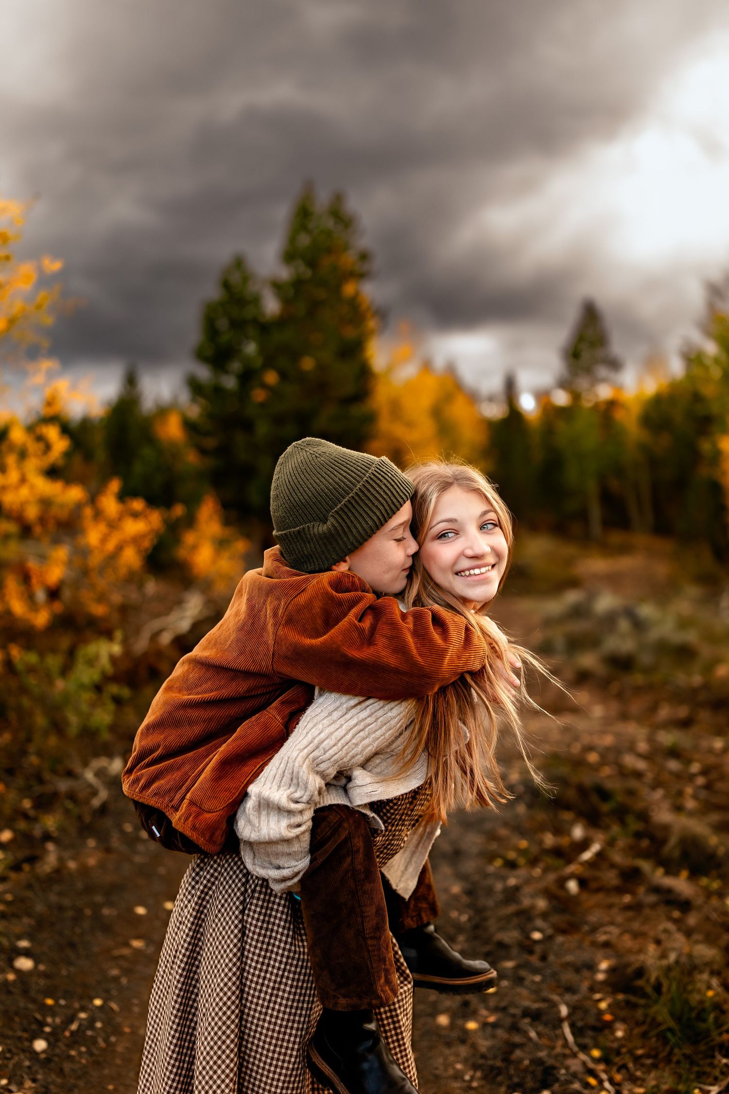 A woman gives a child a piggyback ride outdoors in an autumn forest setting with colorful fall foliage.