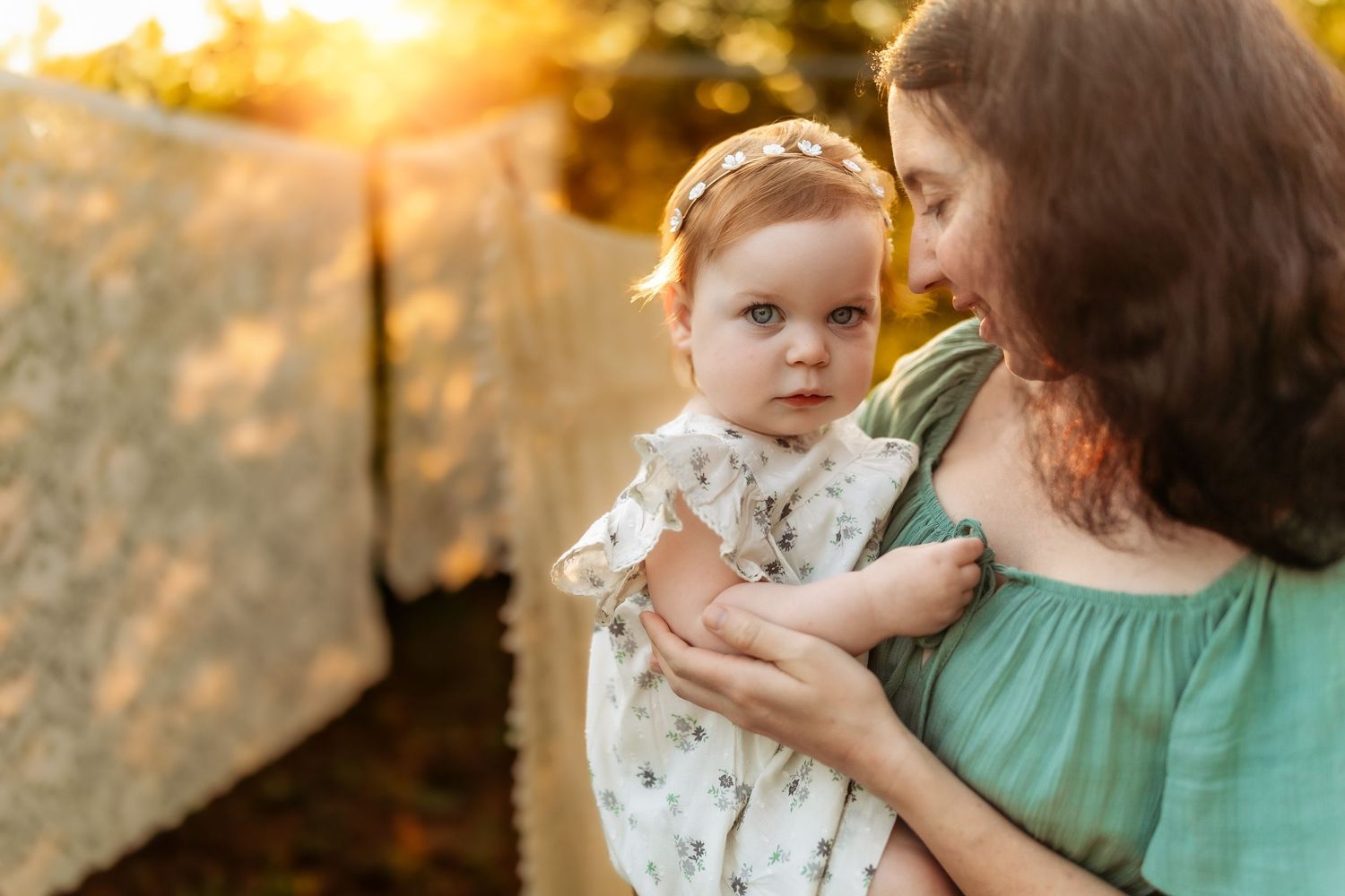 Mother holding baby girl in golden sunset light outdoors, infant in floral dress with headband looks at camera.