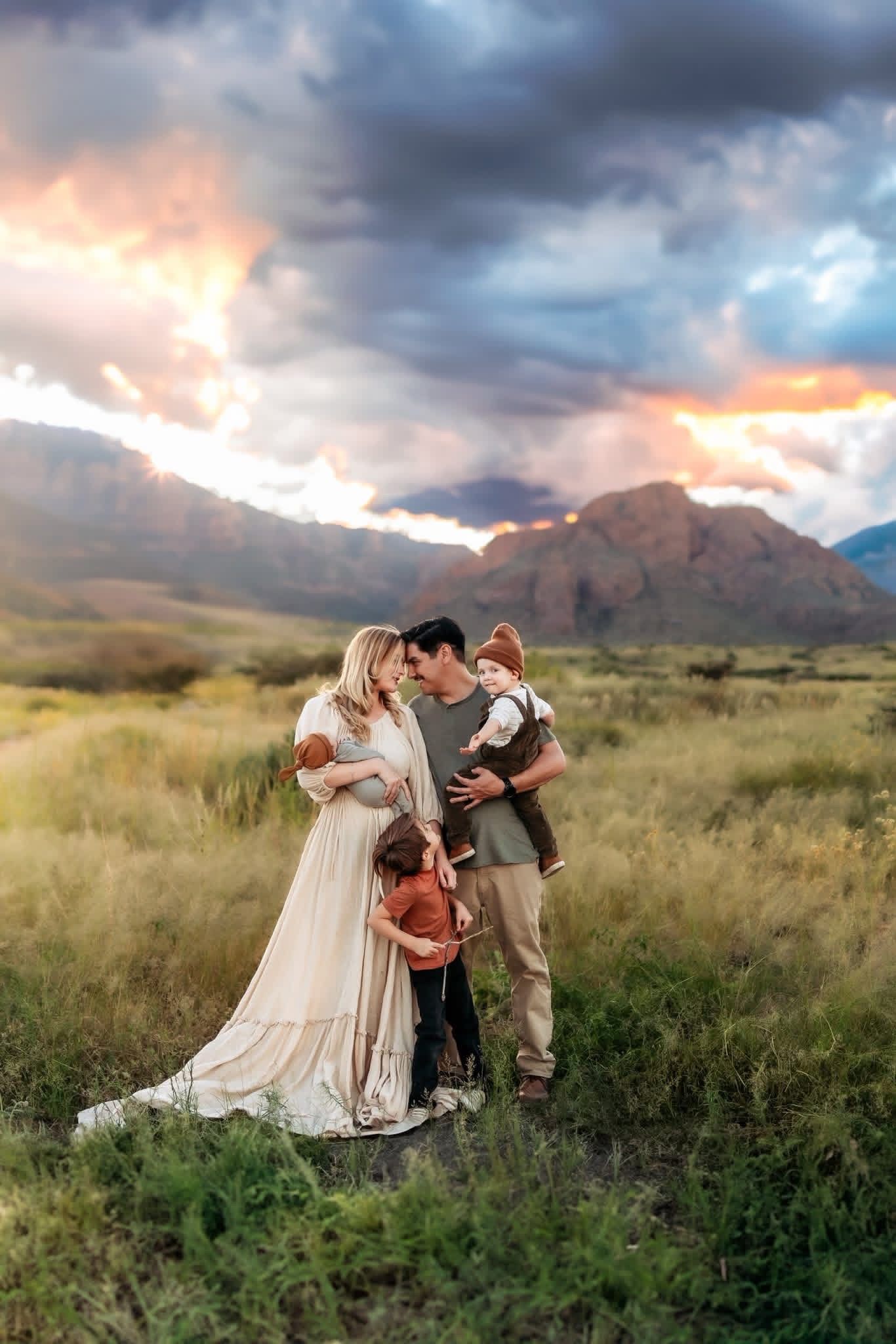 Family of four poses in a mountain meadow at sunset, mother in flowing white dress, dramatic cloudy sky backdrop.