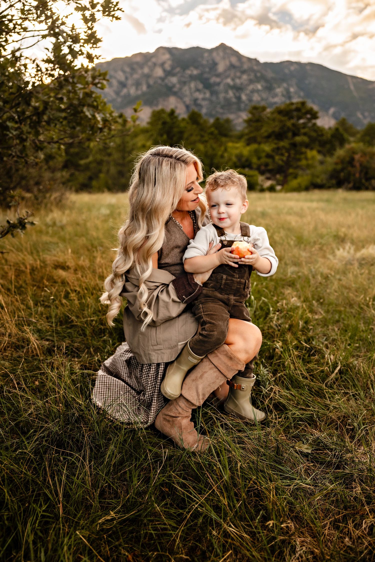 Blonde woman in boots kneels in a grassy field holding a smiling toddler in overalls who holds an apple, with mountains behind.