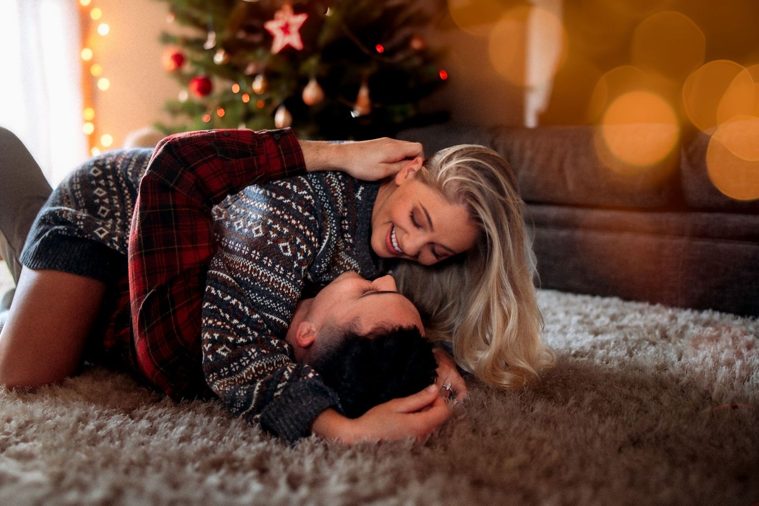Couple cuddling on a carpet in matching Christmas sweaters near a decorated holiday tree with warm bokeh lights.